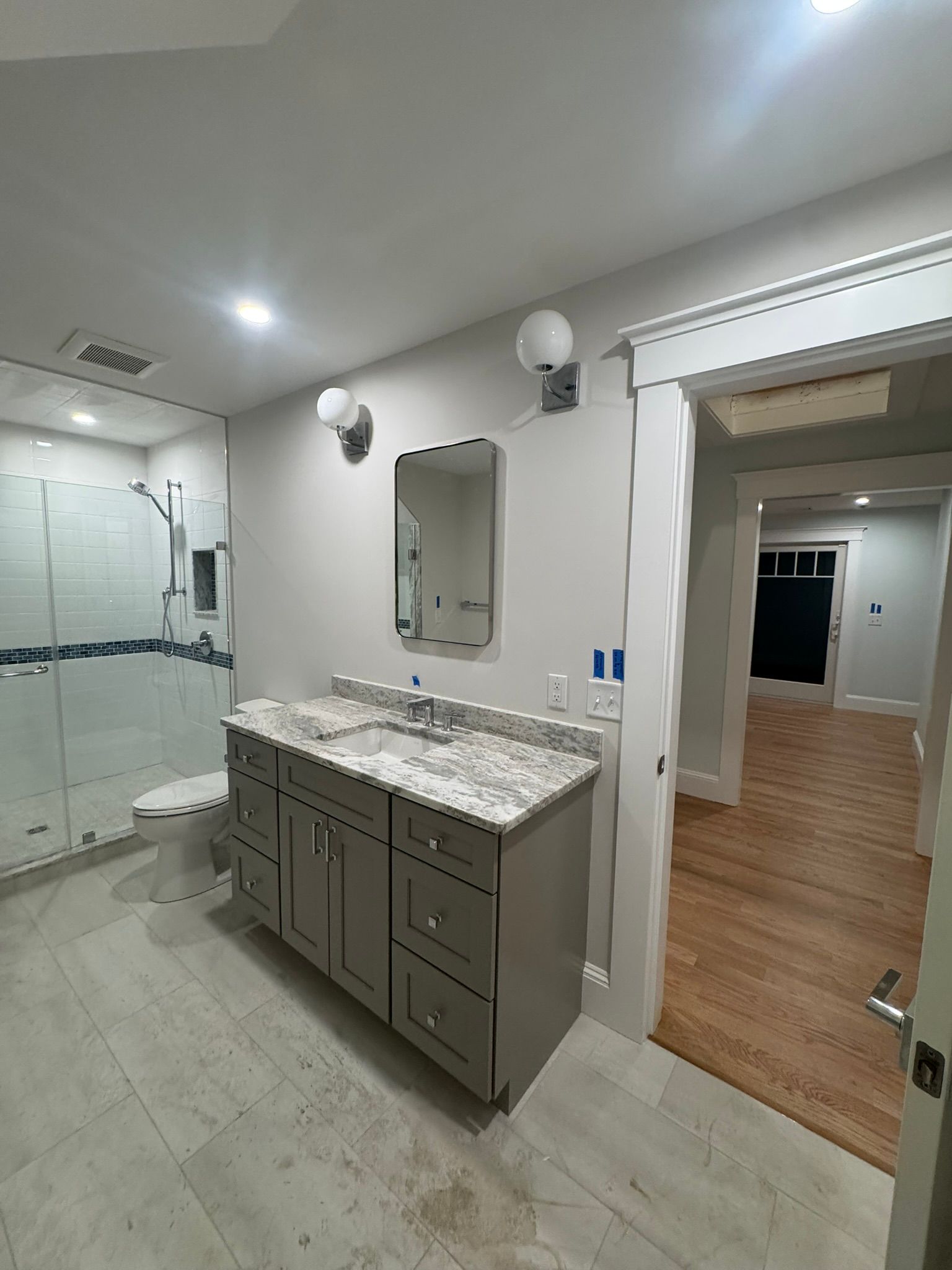 Bathroom vanity with gray cabinets, marble countertop, mirror, and glass shower doors leading to a wood-floored room