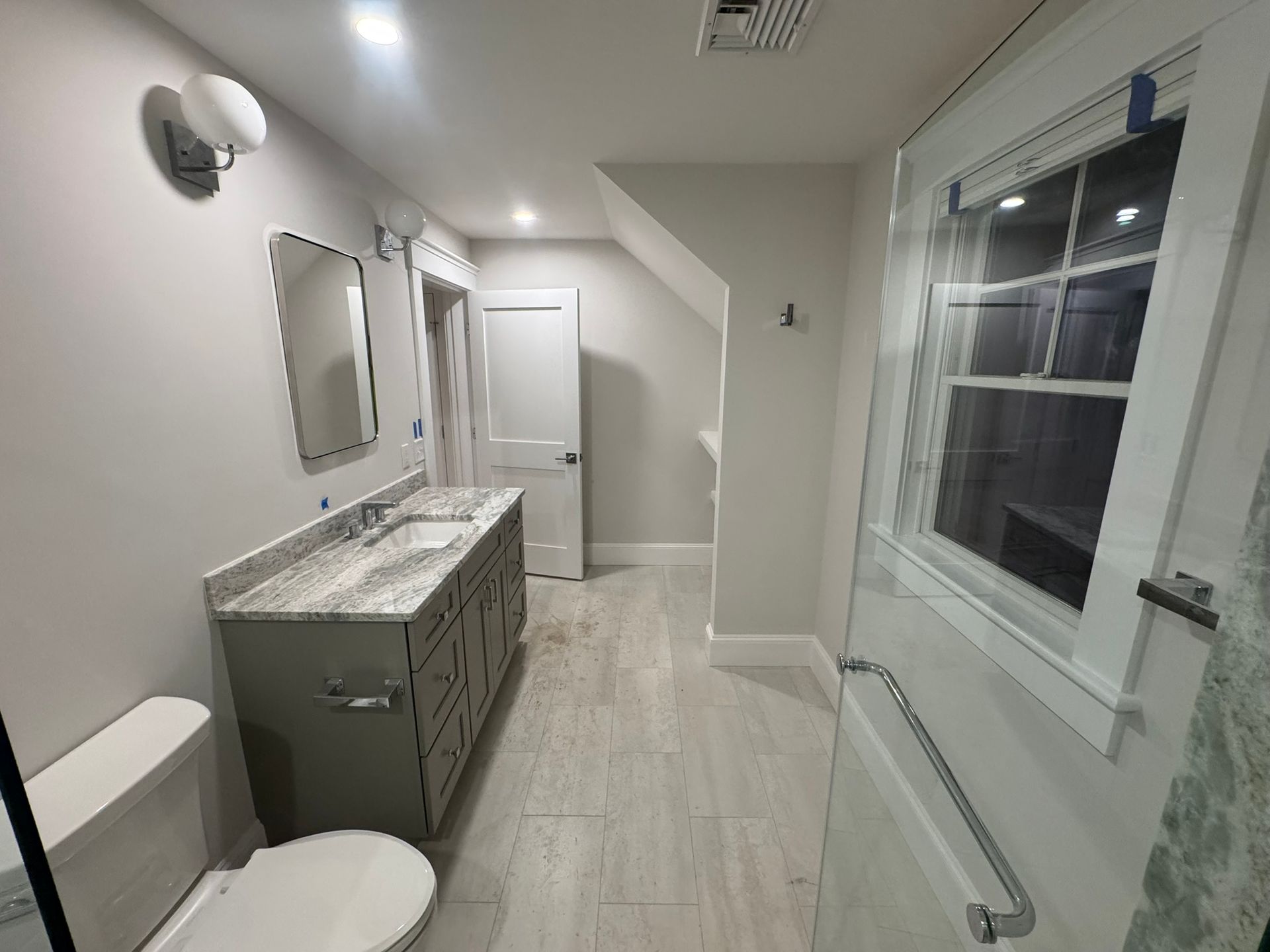 Modern bathroom with double vanity, toilet, large window, and glass shower door; bright white and gray finishes