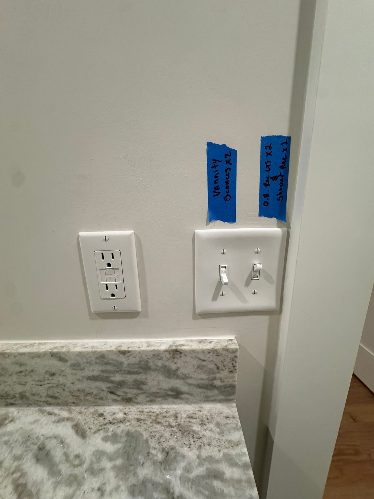 Two wall outlets and a light switch plate with blue tape labels beside a granite countertop.