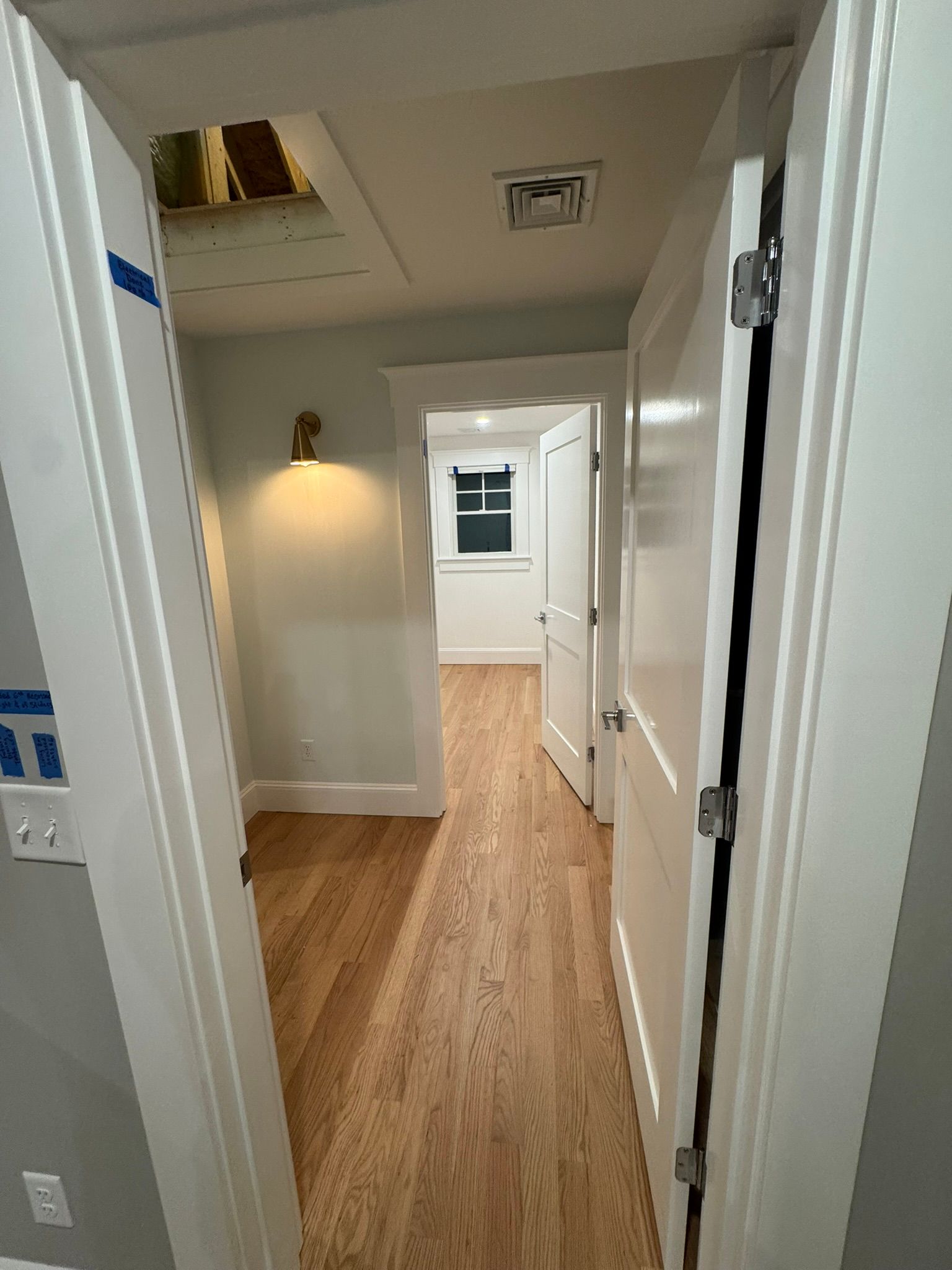Narrow hallway with wood floors, white walls, and a lit wall sconce leading to a bright room.