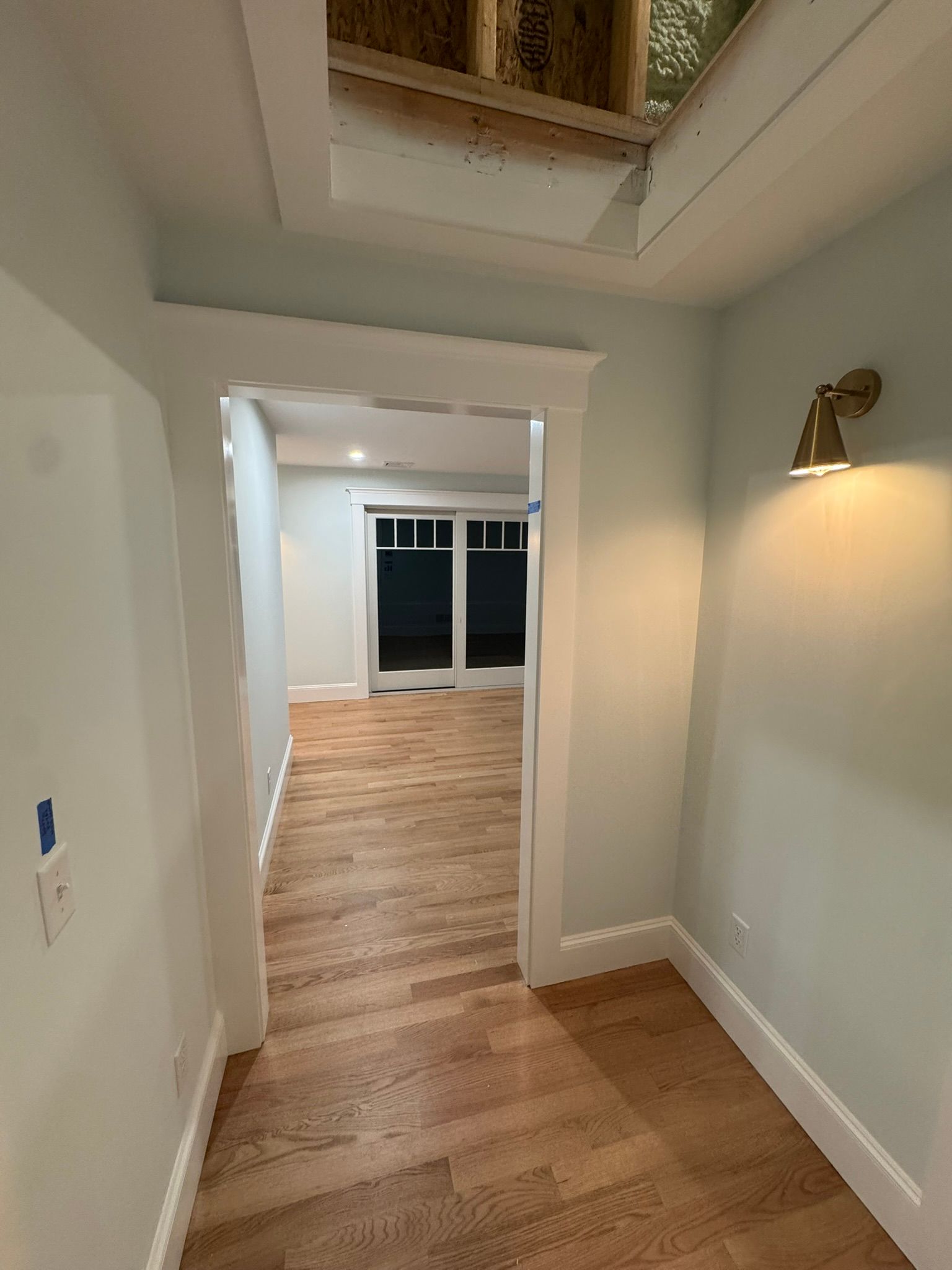 Narrow hallway with hardwood floors, white walls, wall sconce, and a doorway opening to a bright room with glass doors