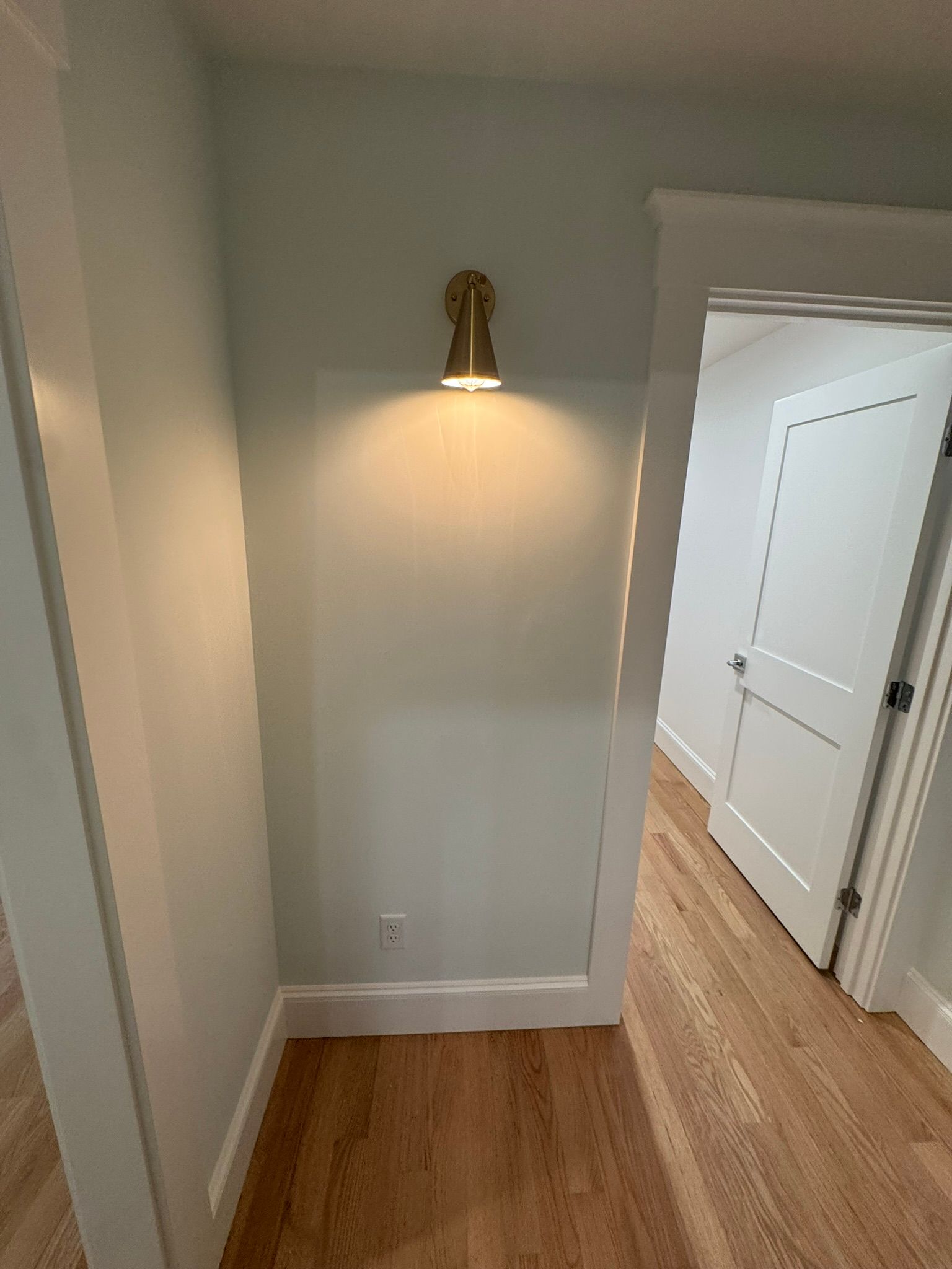 Empty hallway with hardwood floors, gray walls, and a lit wall sconce near a white doorway.