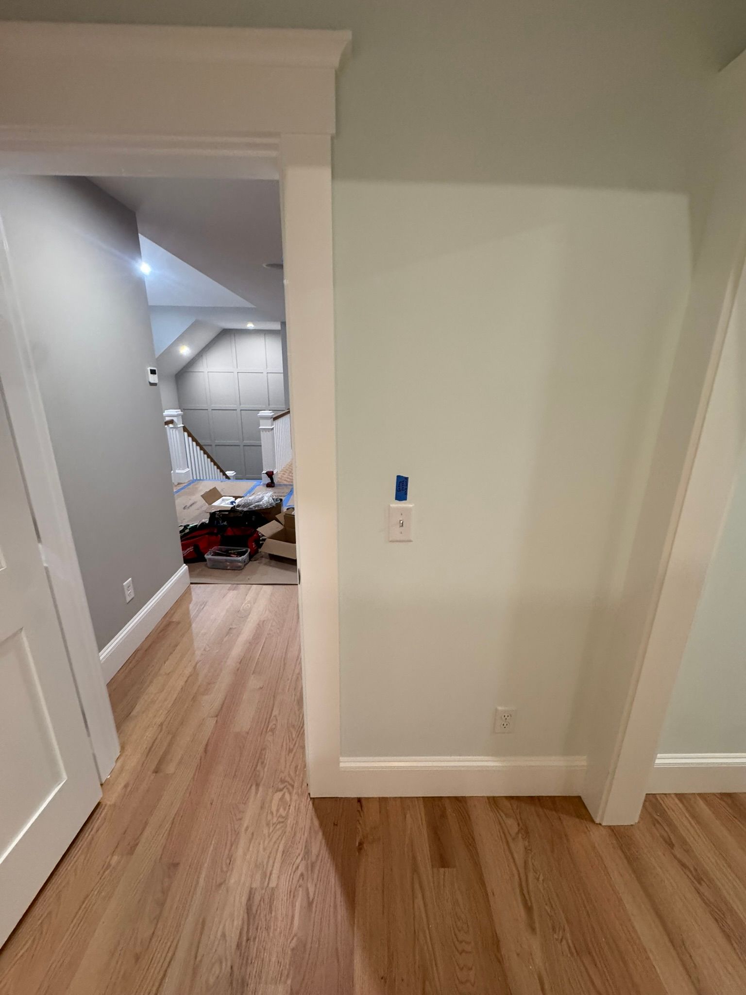 Hallway with wood floors leading to a cluttered room, white walls, and a doorway on the left.