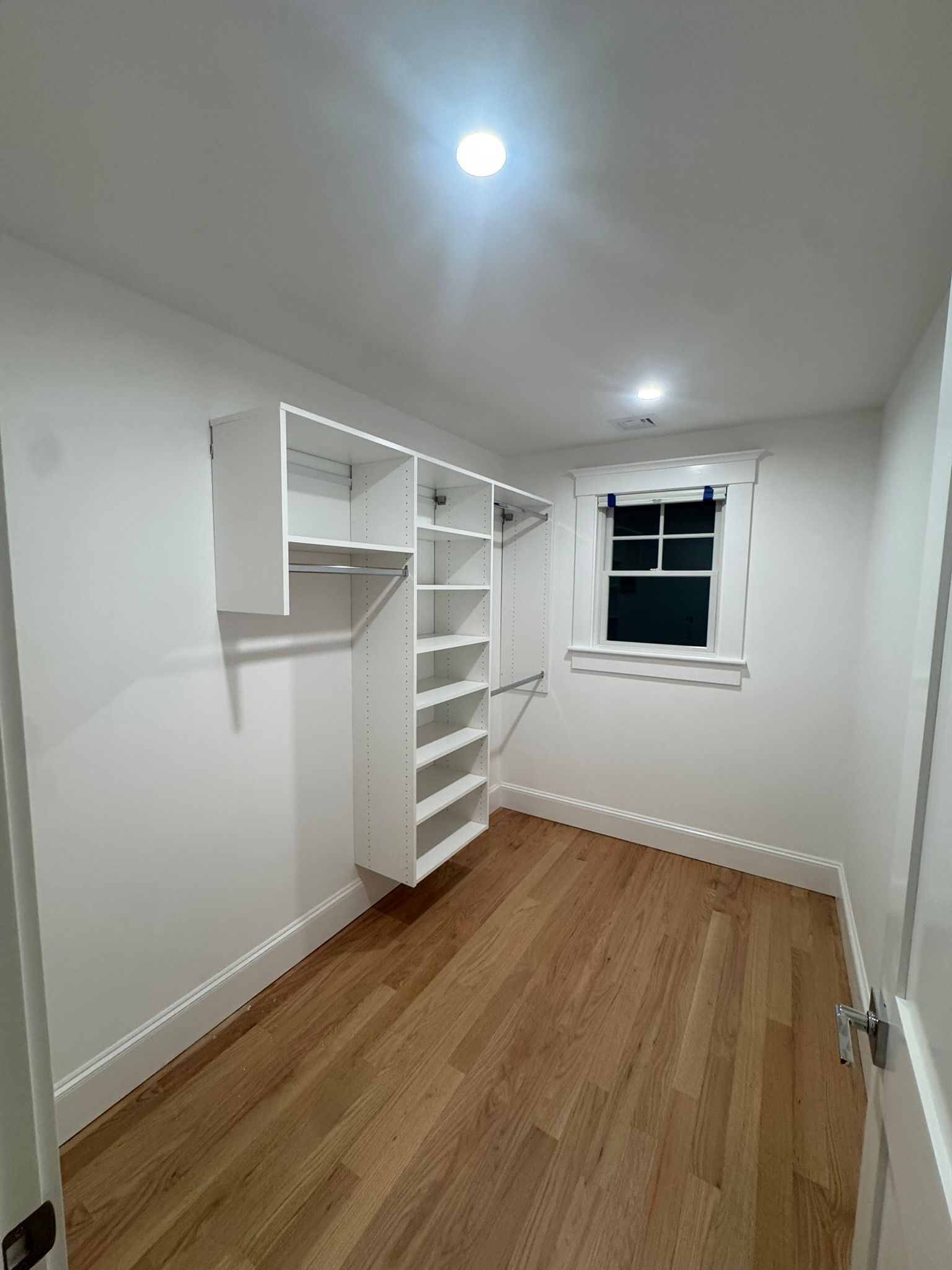 Empty white bedroom with wood floor, built-in shelving, and a window.