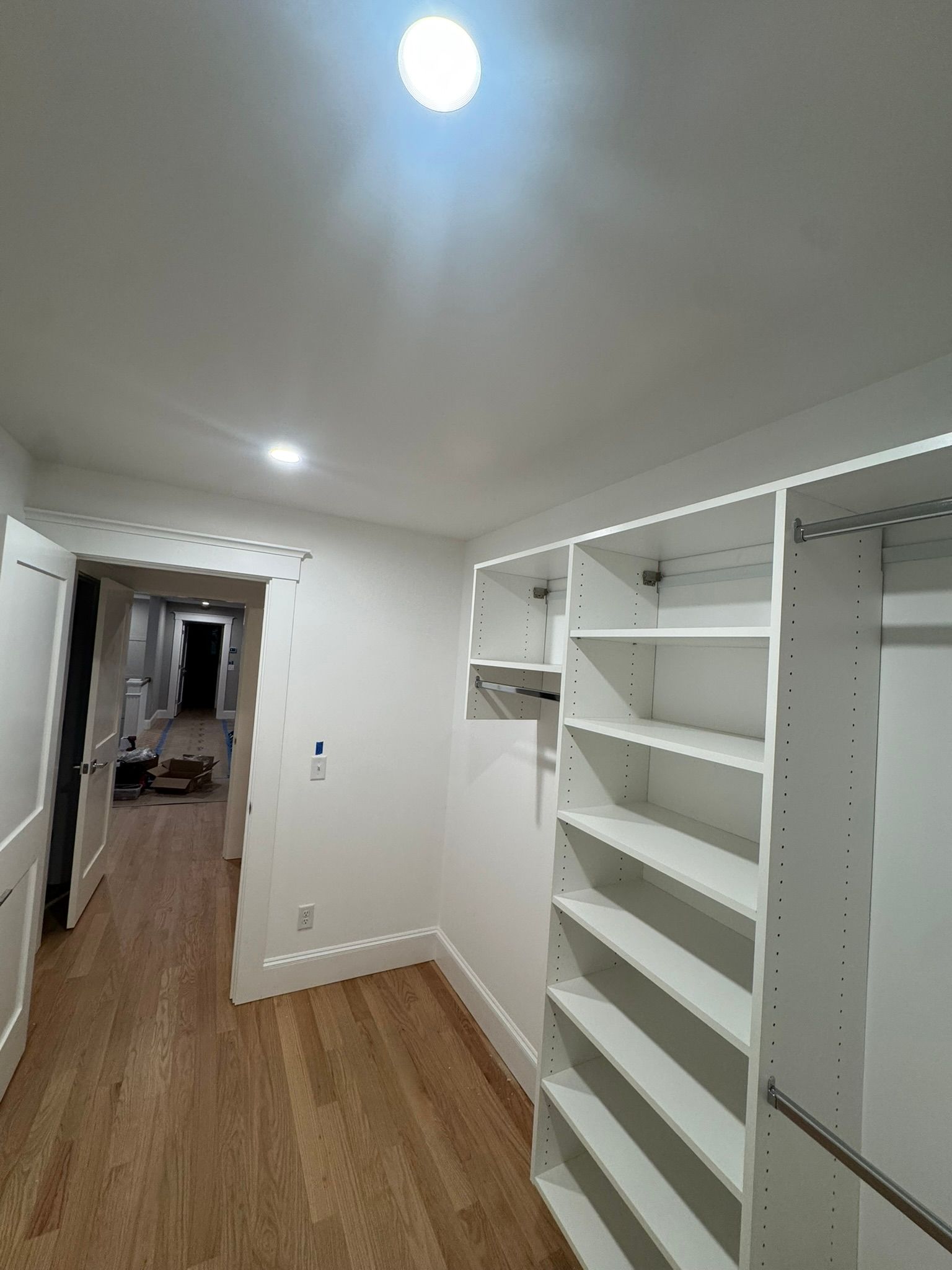 Narrow white closet or pantry with built-in shelves, wood floor, and ceiling light in a hallway