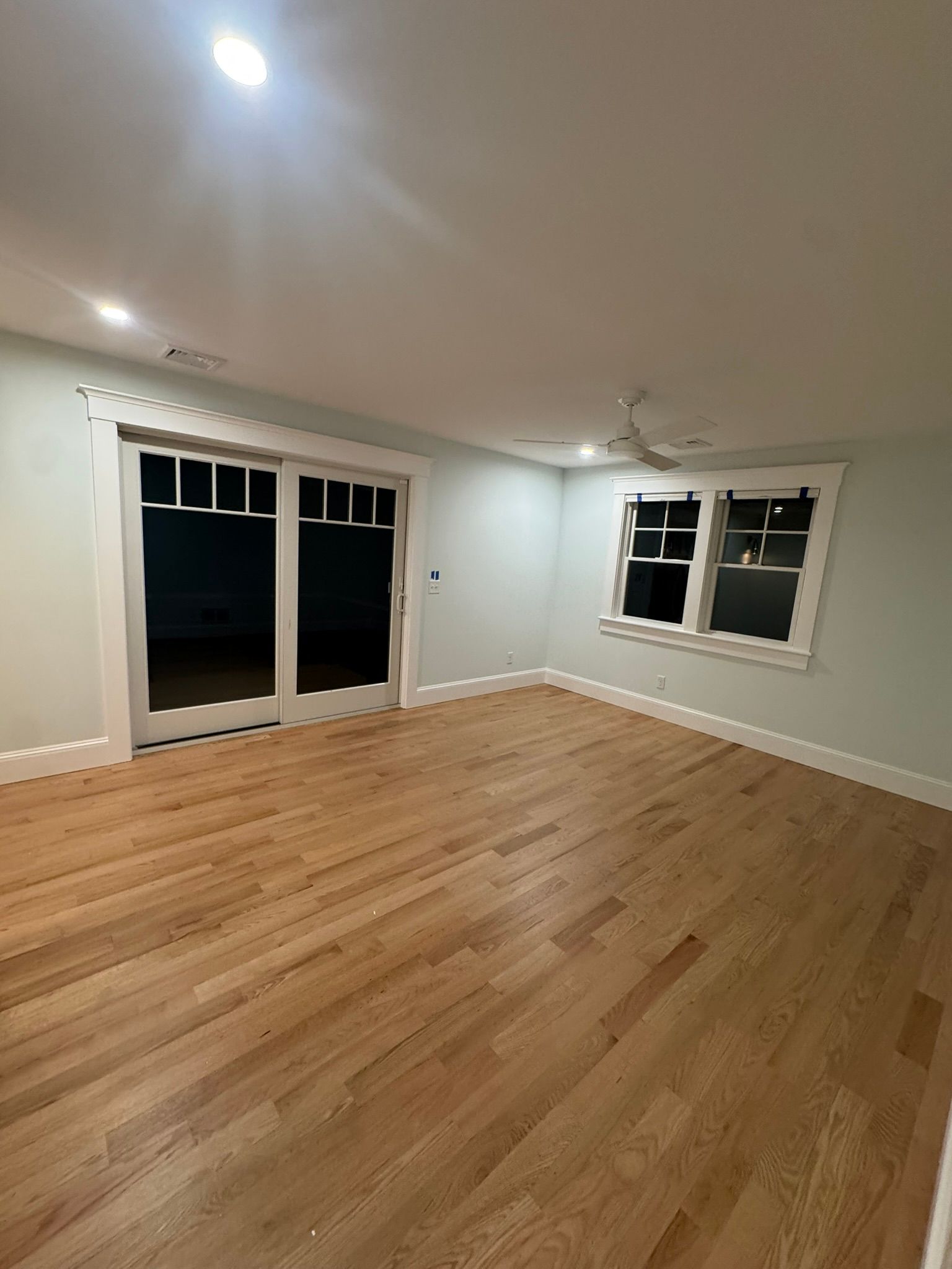 Empty living room with hardwood floors, white walls, ceiling fan, and large windows and glass doors at night