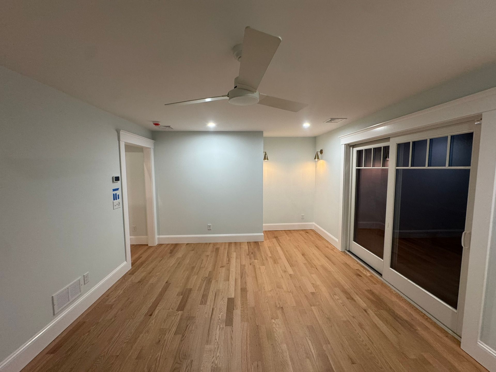 Empty modern living room with wood floors, ceiling fan, white walls, and sliding glass doors to a deck.