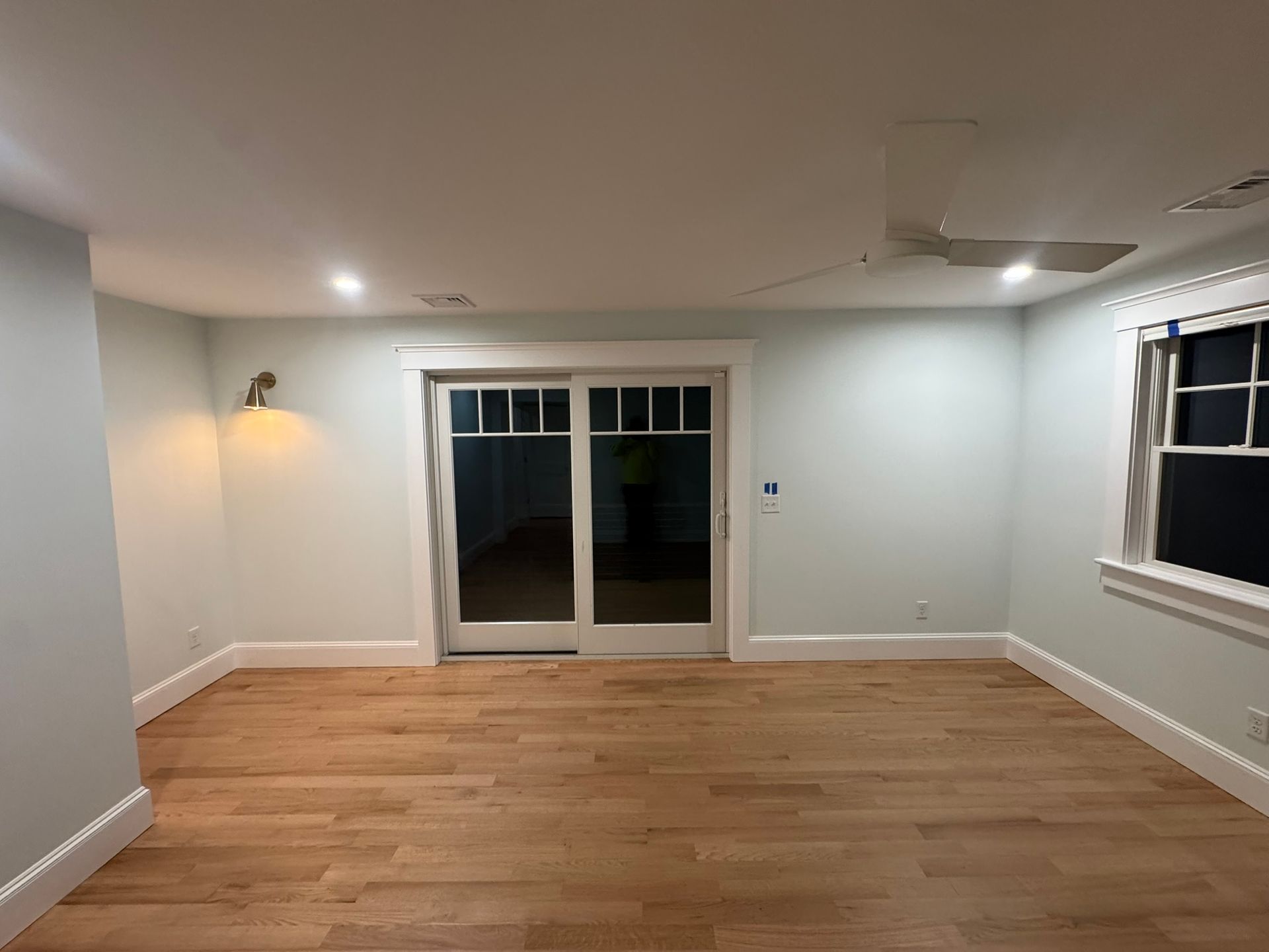 Empty living room with wood floors, white walls, ceiling lights, and sliding glass doors to a dark patio.