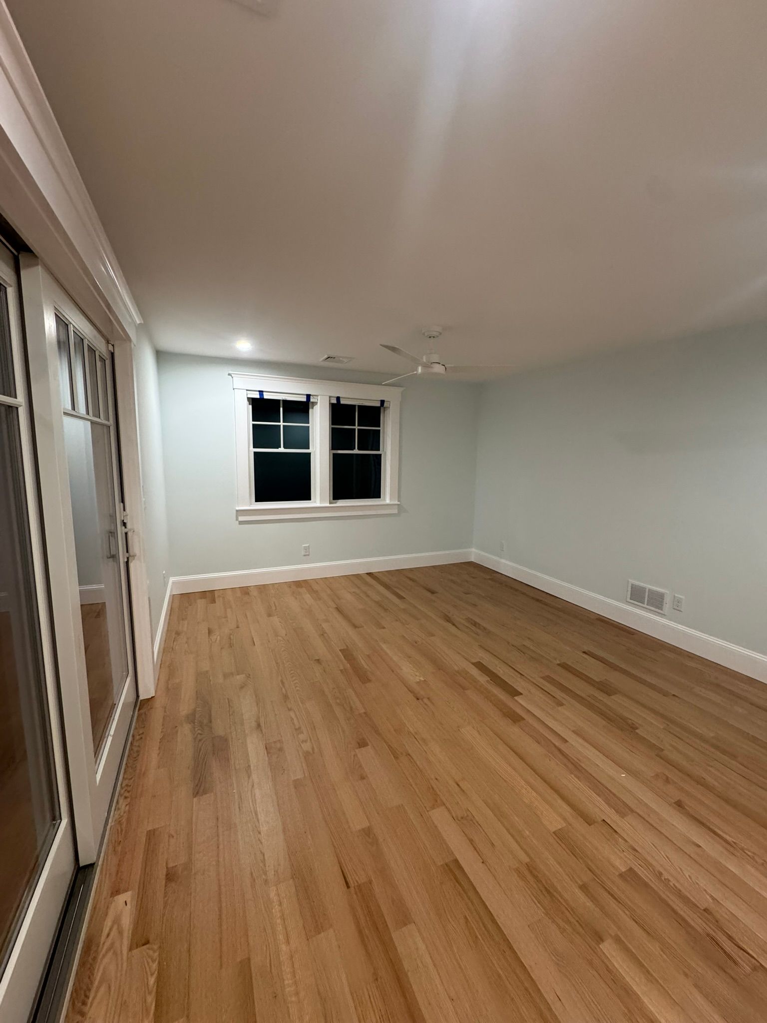 Empty light-gray room with hardwood floors, recessed ceiling lights, and a window on the far wall.