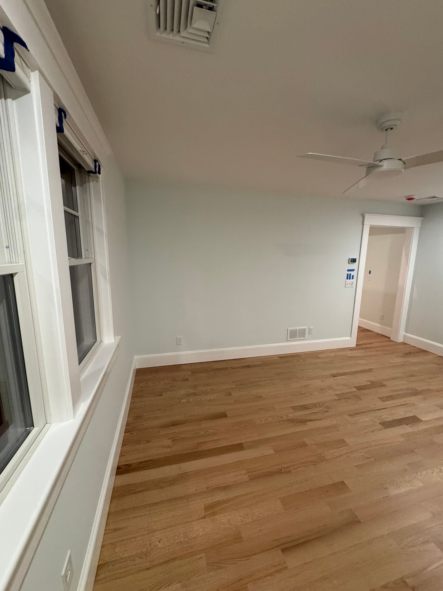 Empty room with hardwood floors, white walls, a window, ceiling fan, and open doorway