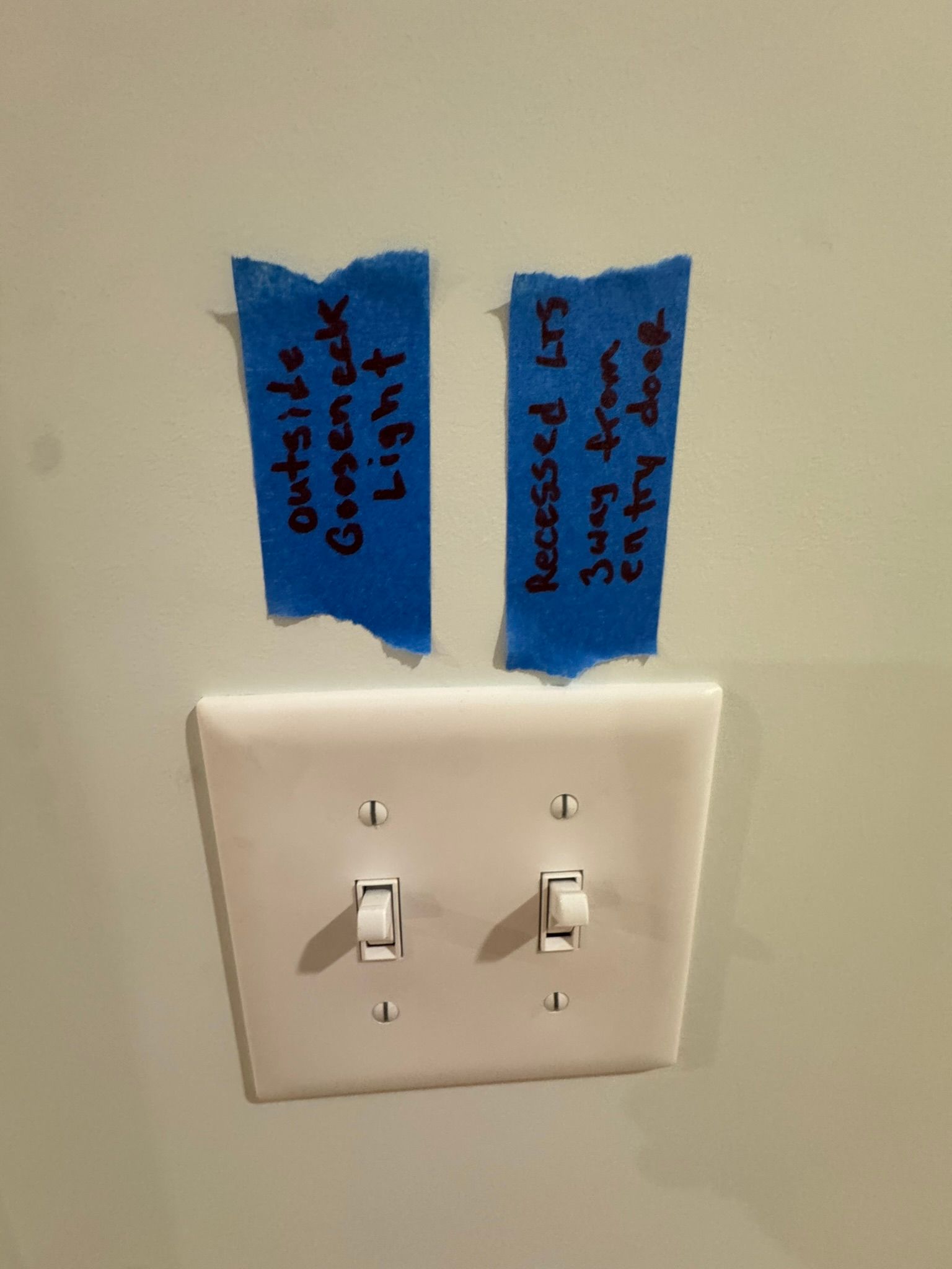 Light switch plate with two switches labeled by blue tape notes on a wall