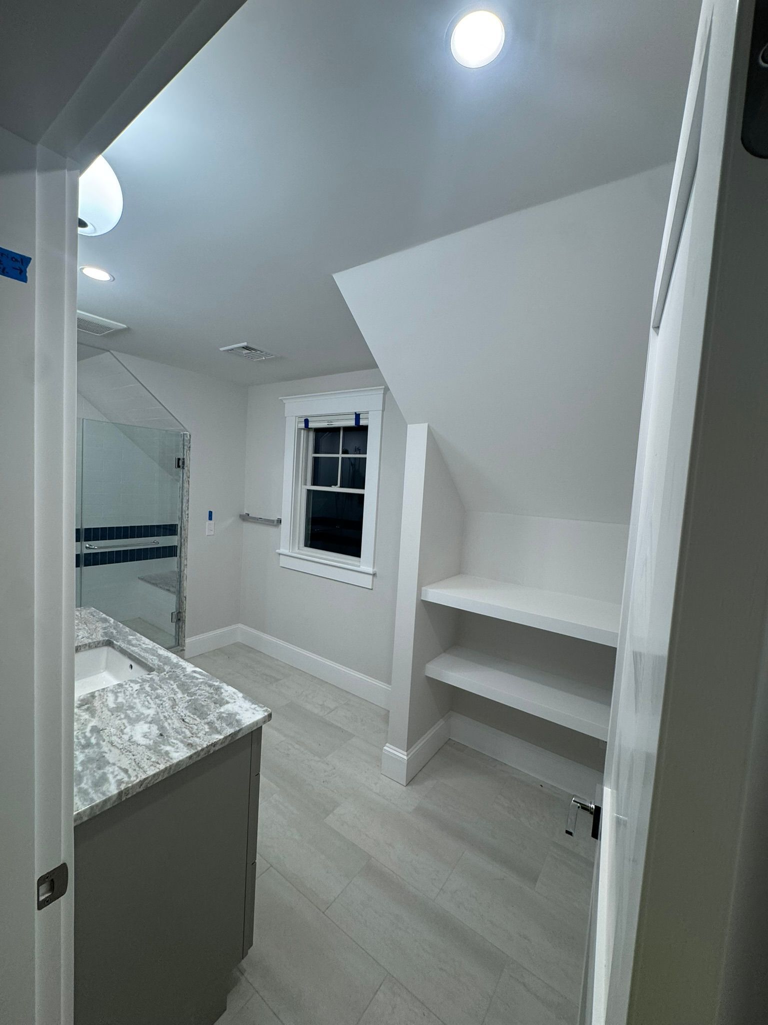 Small white bathroom with slanted ceiling, window, vanity, and open shelving.