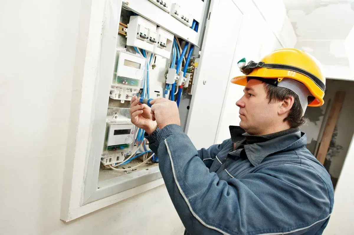 Electrician in yellow hard hat working inside an open wall electrical panel, adjusting wires.