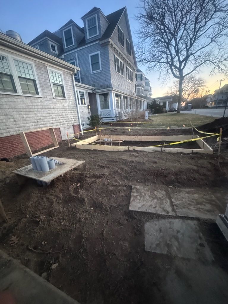 Muddy construction site beside a gray house, with excavated ground and yellow caution tape at dusk