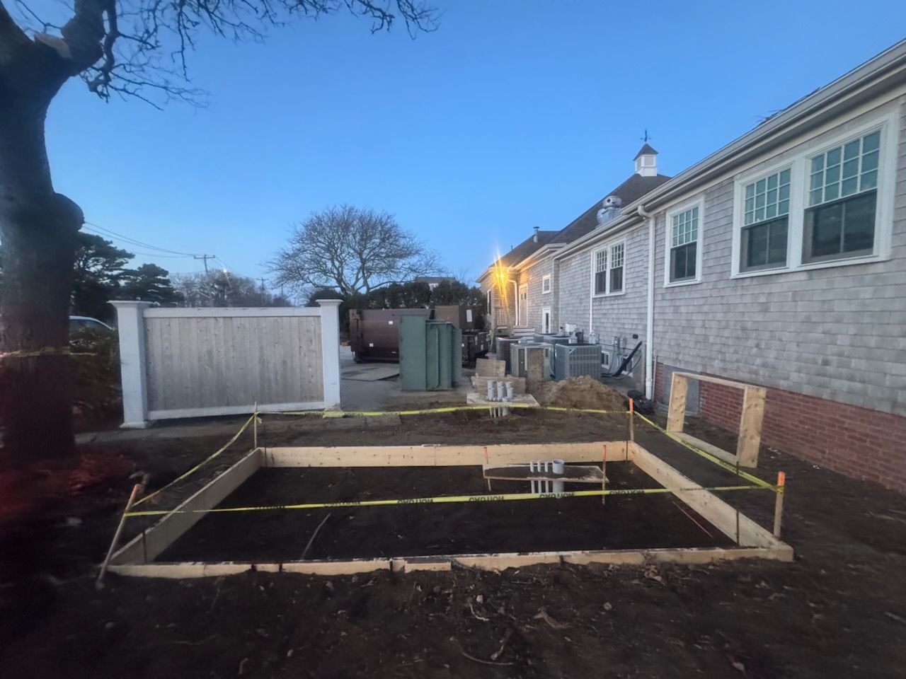 Construction site with wooden foundation framing beside a white brick building at dusk