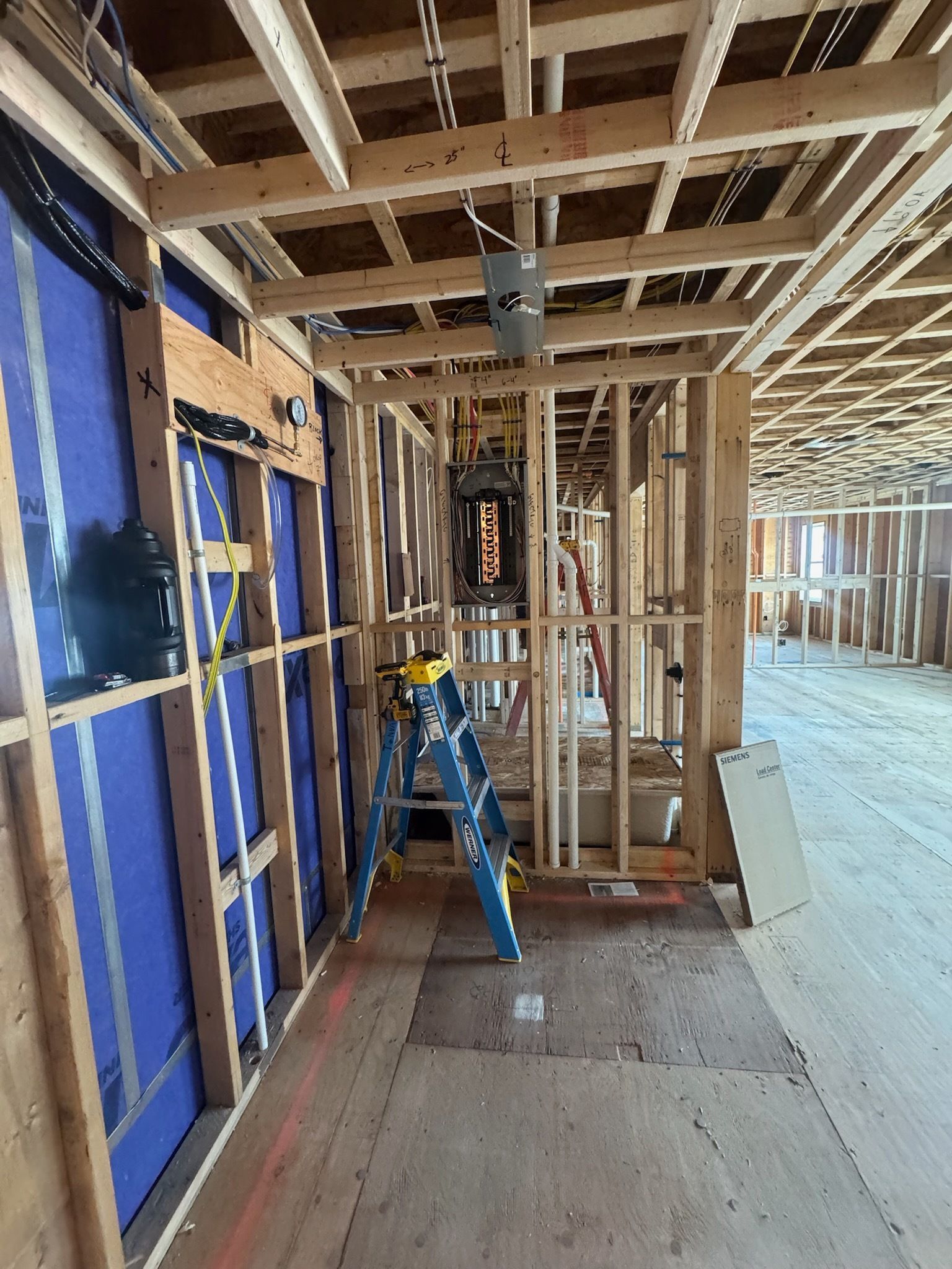 Interior of a house under construction, with exposed framing, blue sheathing, and a ladder in a hallway.
