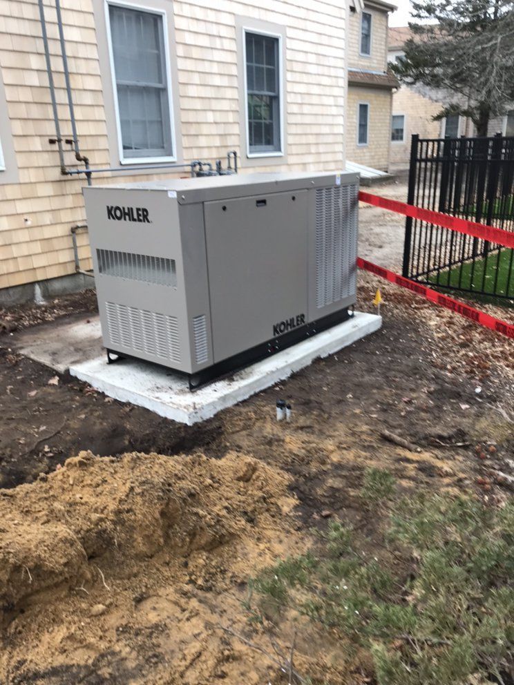 Gray Kohler generator on a concrete pad beside a beige house, with dirt and a red safety fence nearby.