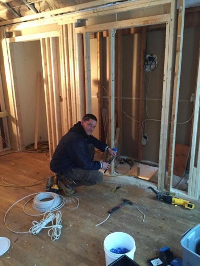 Worker kneels in a framed room, using a power tool amid wiring and construction materials.