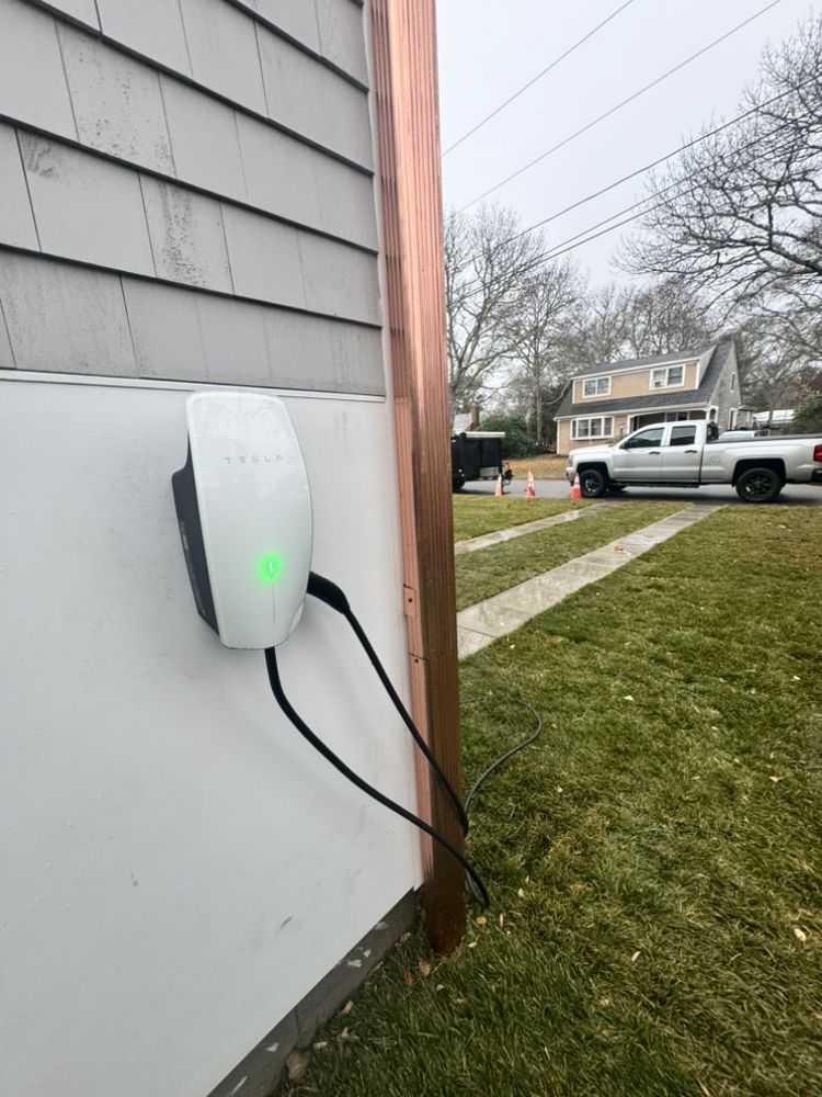Wall-mounted EV charger with glowing green light beside a house in a residential yard.