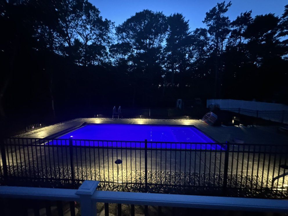 Backyard swimming pool glowing blue at dusk, surrounded by fence and trees