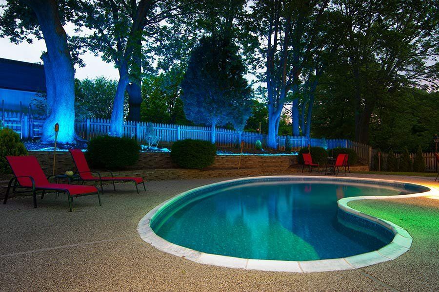 Backyard pool at dusk with blue-lit trees and red lounge chairs