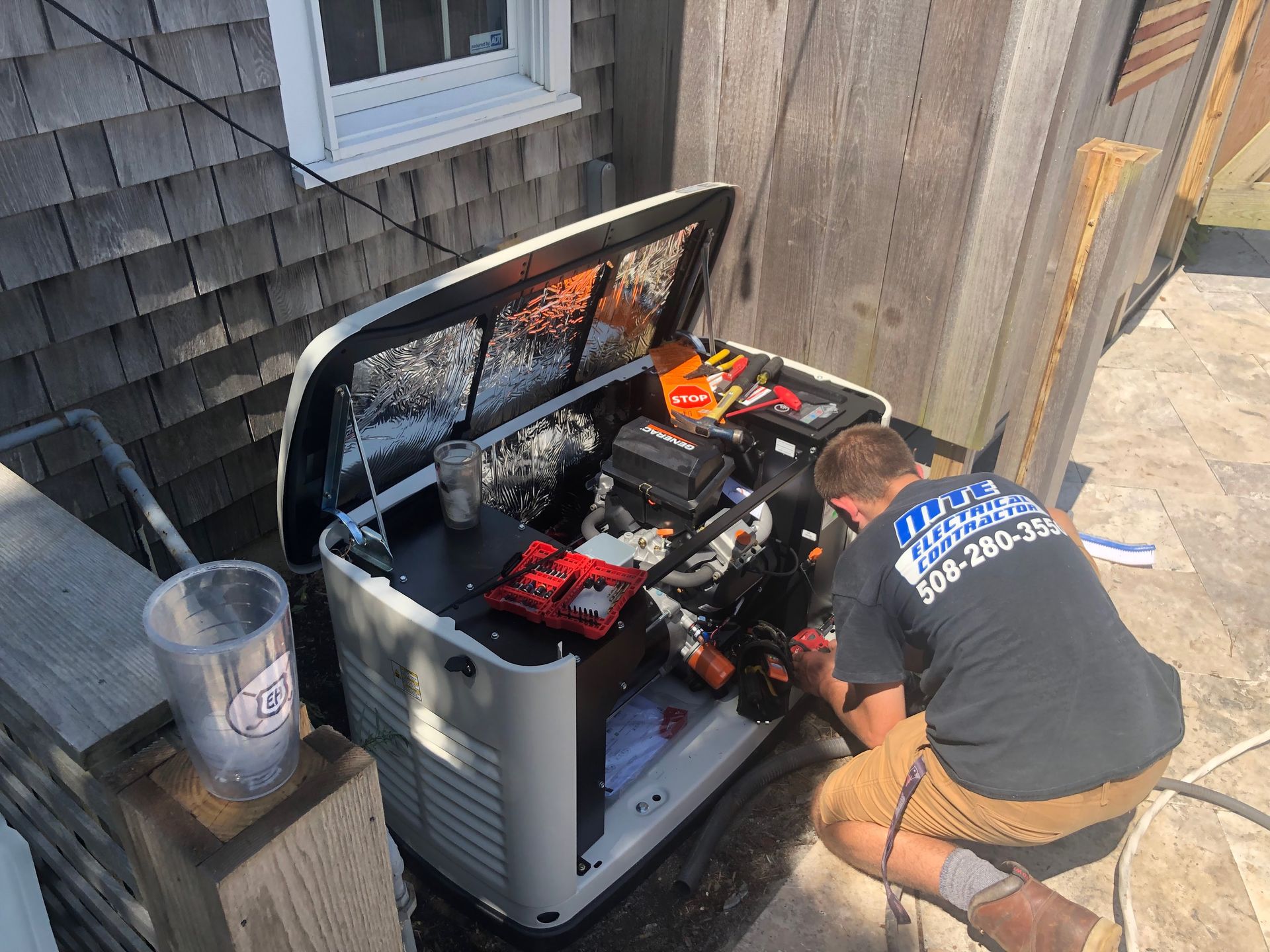 Person kneeling by open generator, inspecting engine on patio beside house and fence.