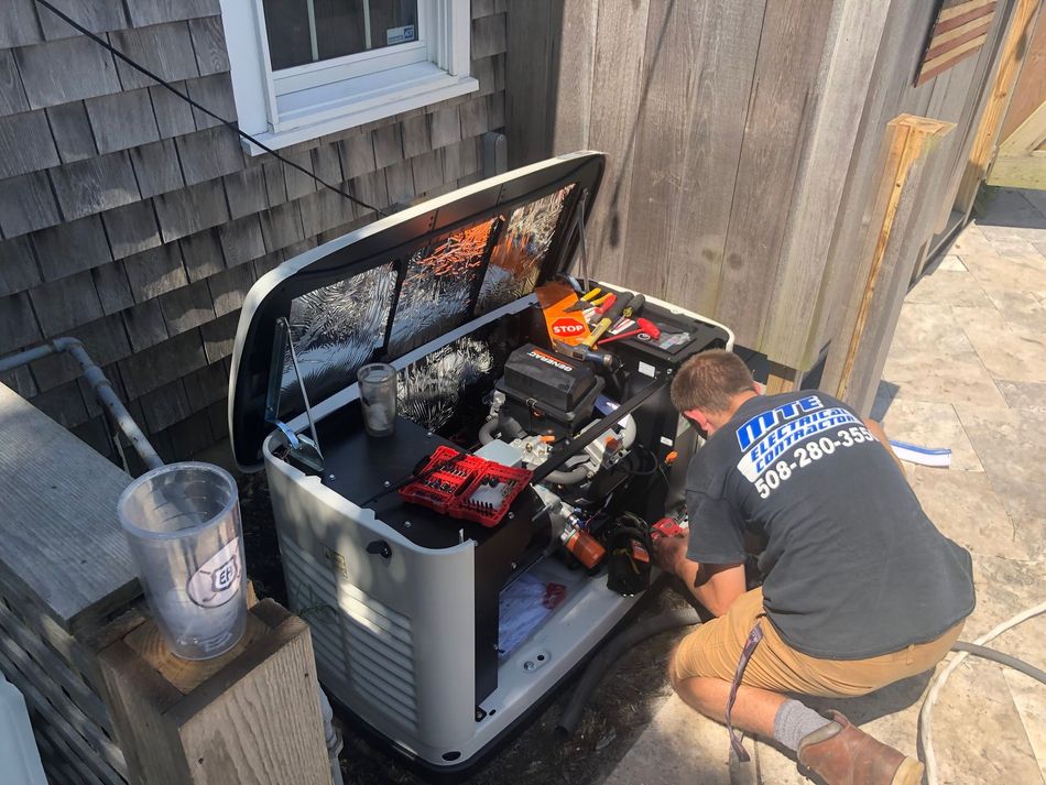 Person kneeling beside an open generator, working in a narrow outdoor alley next to a wooden fence and house.