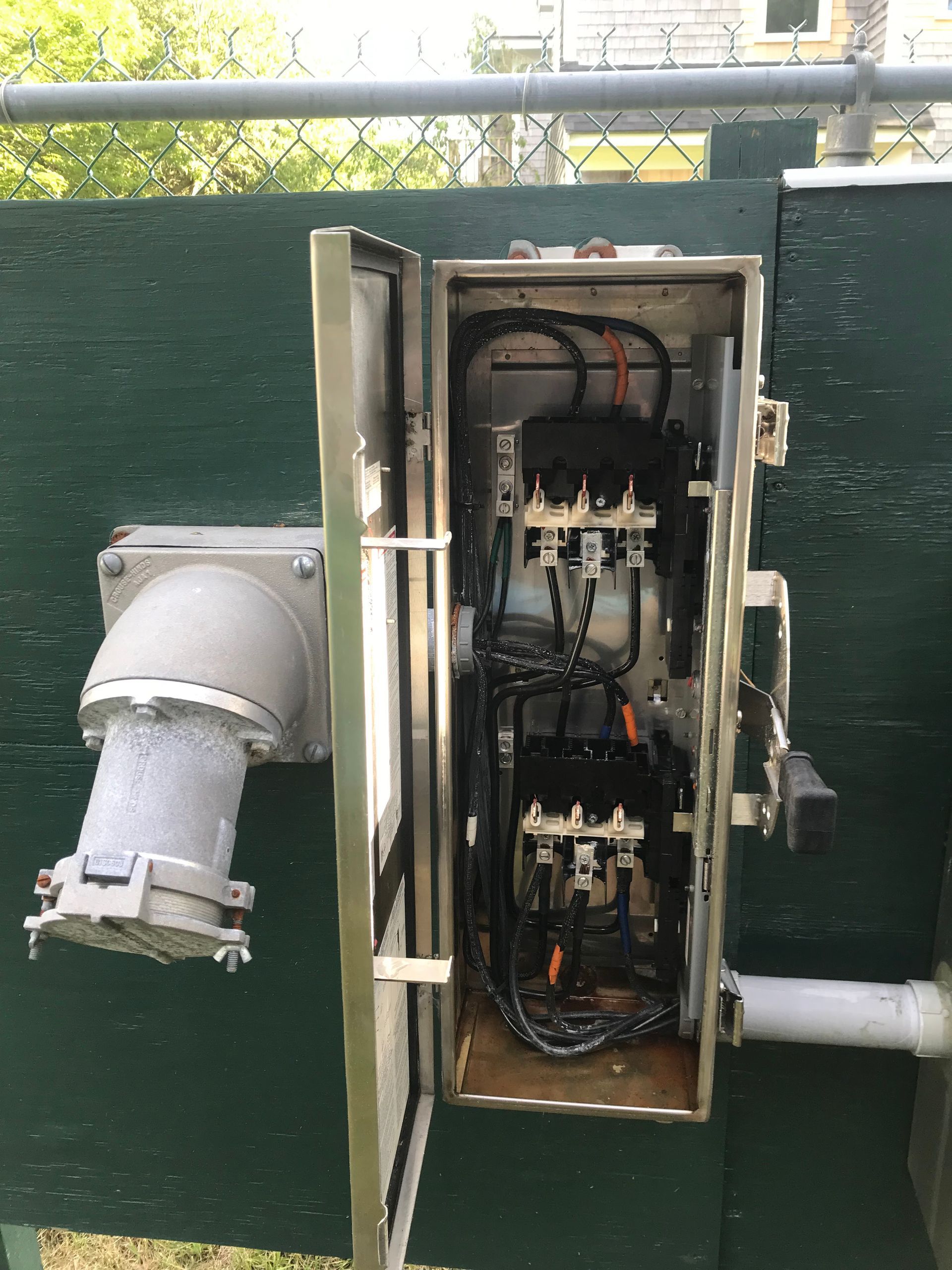 Open electrical control panel mounted on a green wall with conduit pipes and wiring visible inside