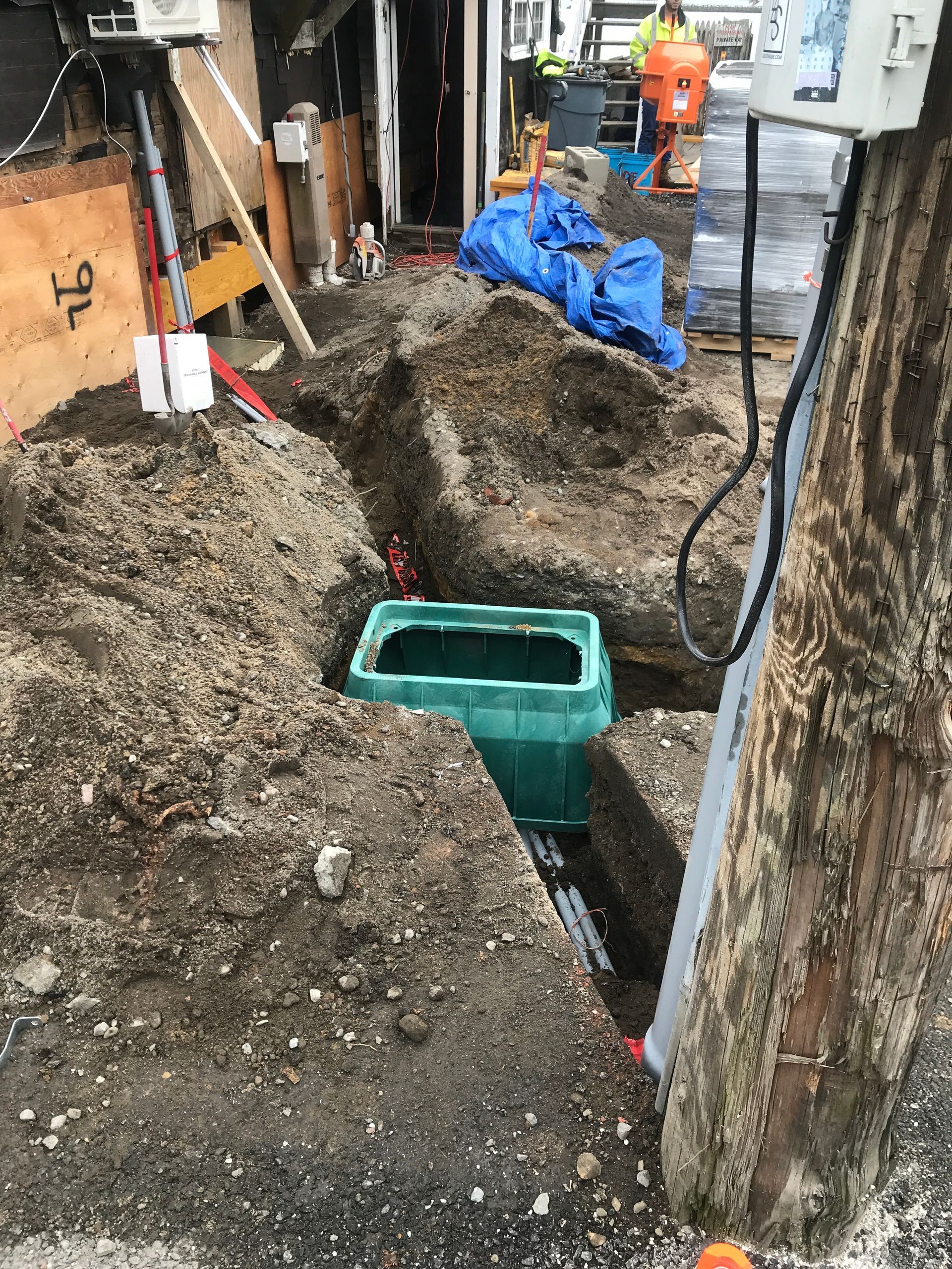 Mud-filled trench with a green utility box and tools at a construction site, with workers in the background