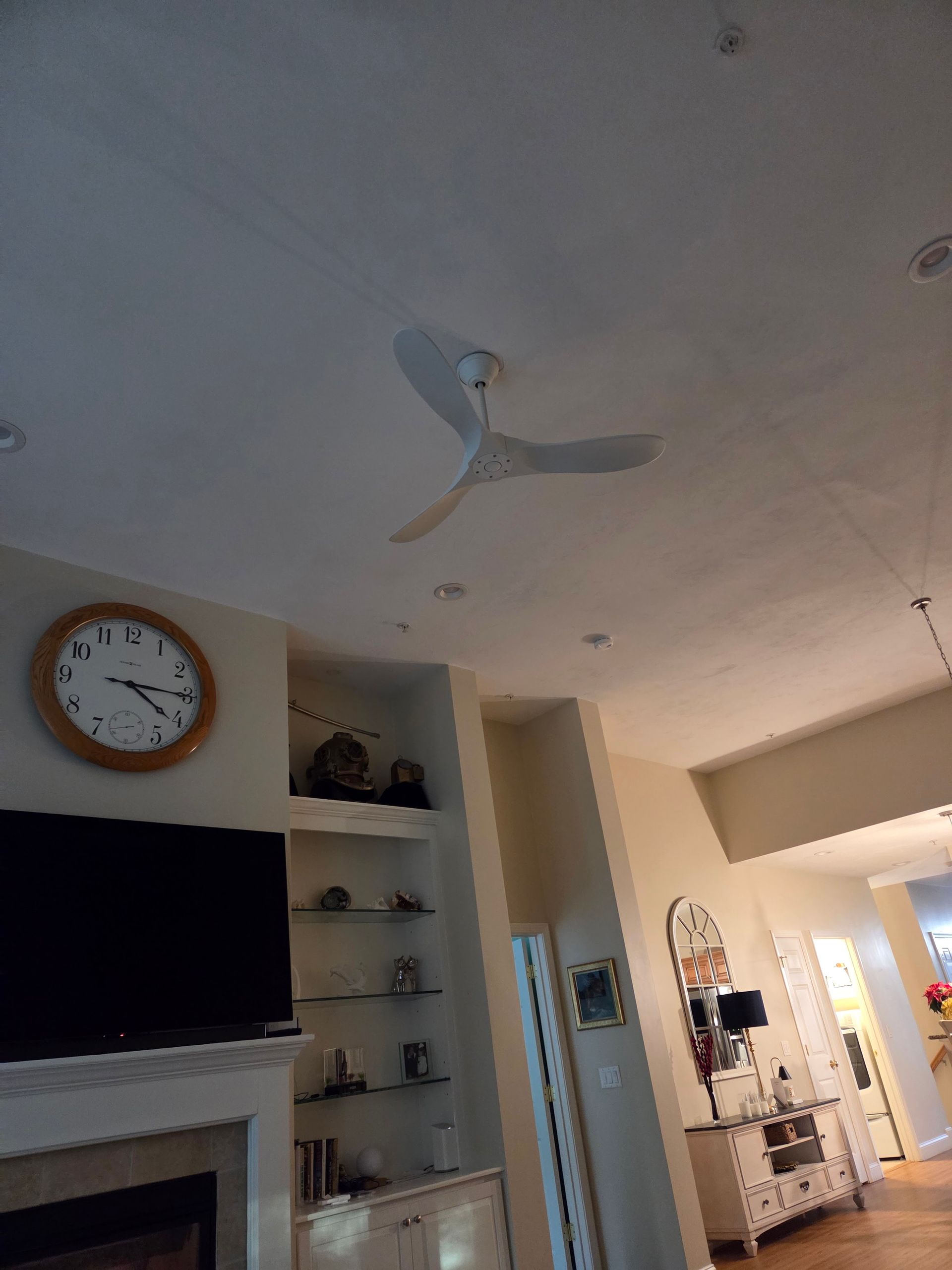 Living room with a ceiling fan, wall clock, TV, shelves, and a hallway in warm lighting