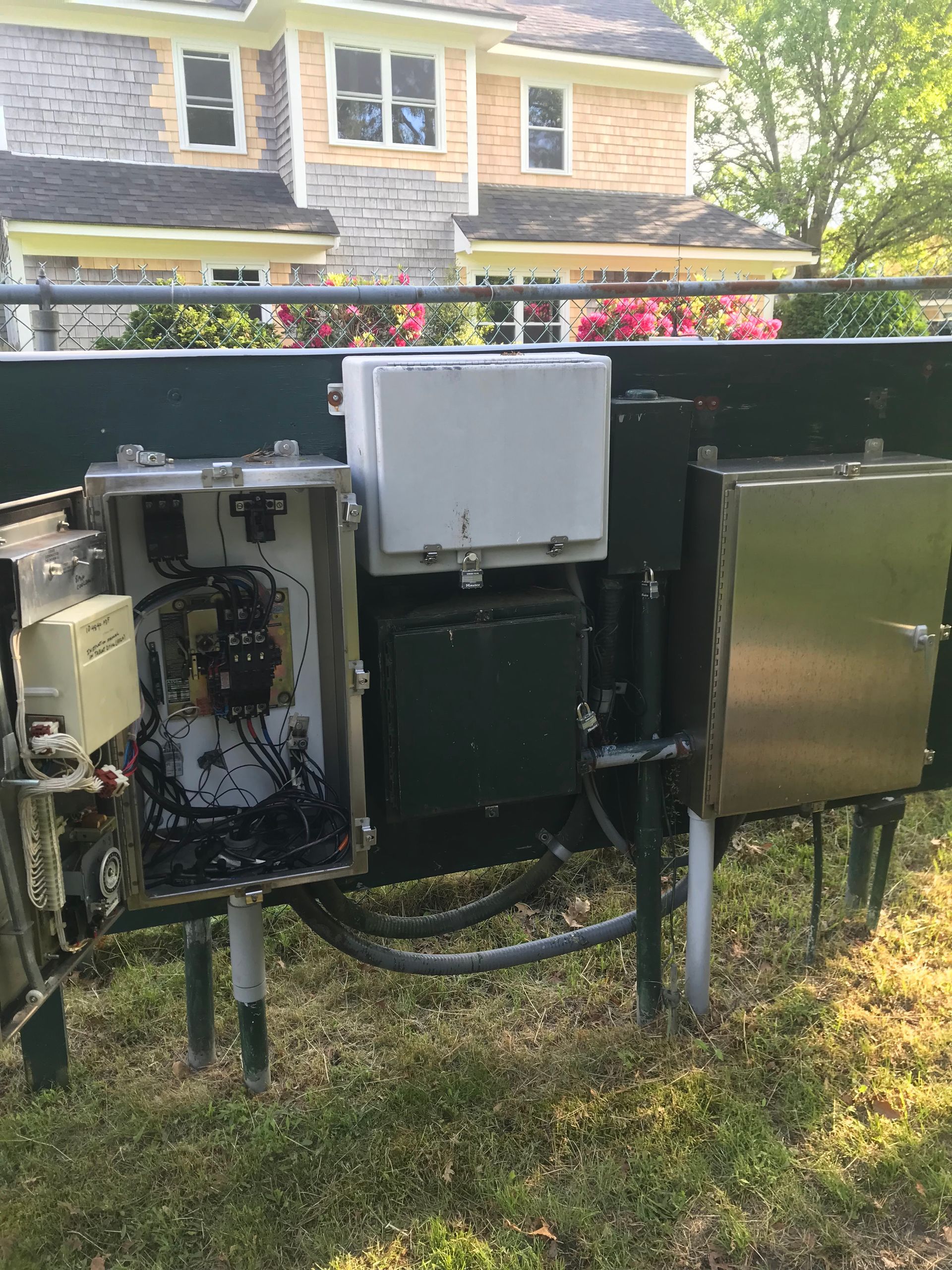 Open electrical and utility boxes mounted outdoors by a house, with exposed wiring and grass below