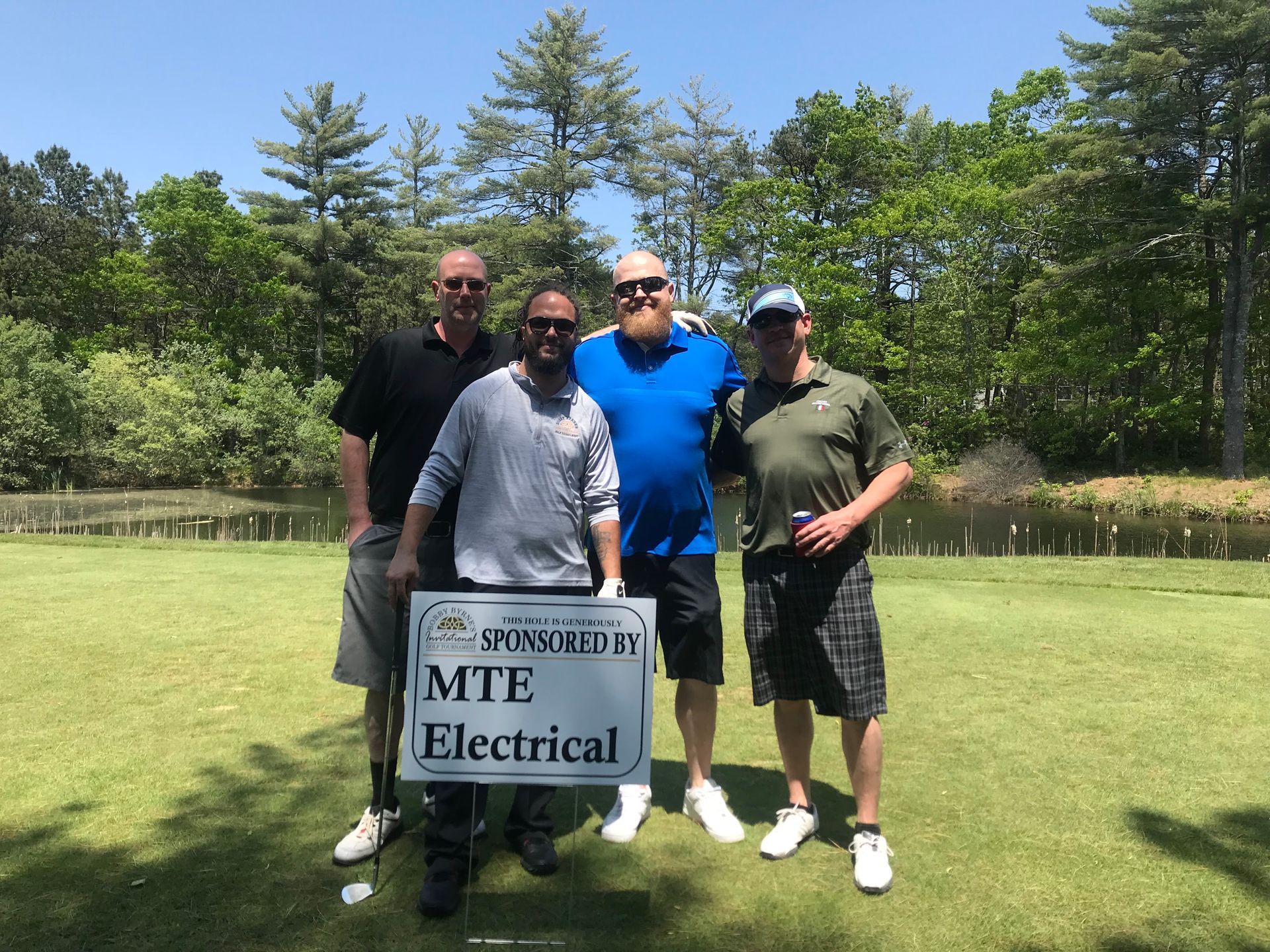 Four people posing on a lawn beside an “MTE Electrical” sign, with trees in the background.