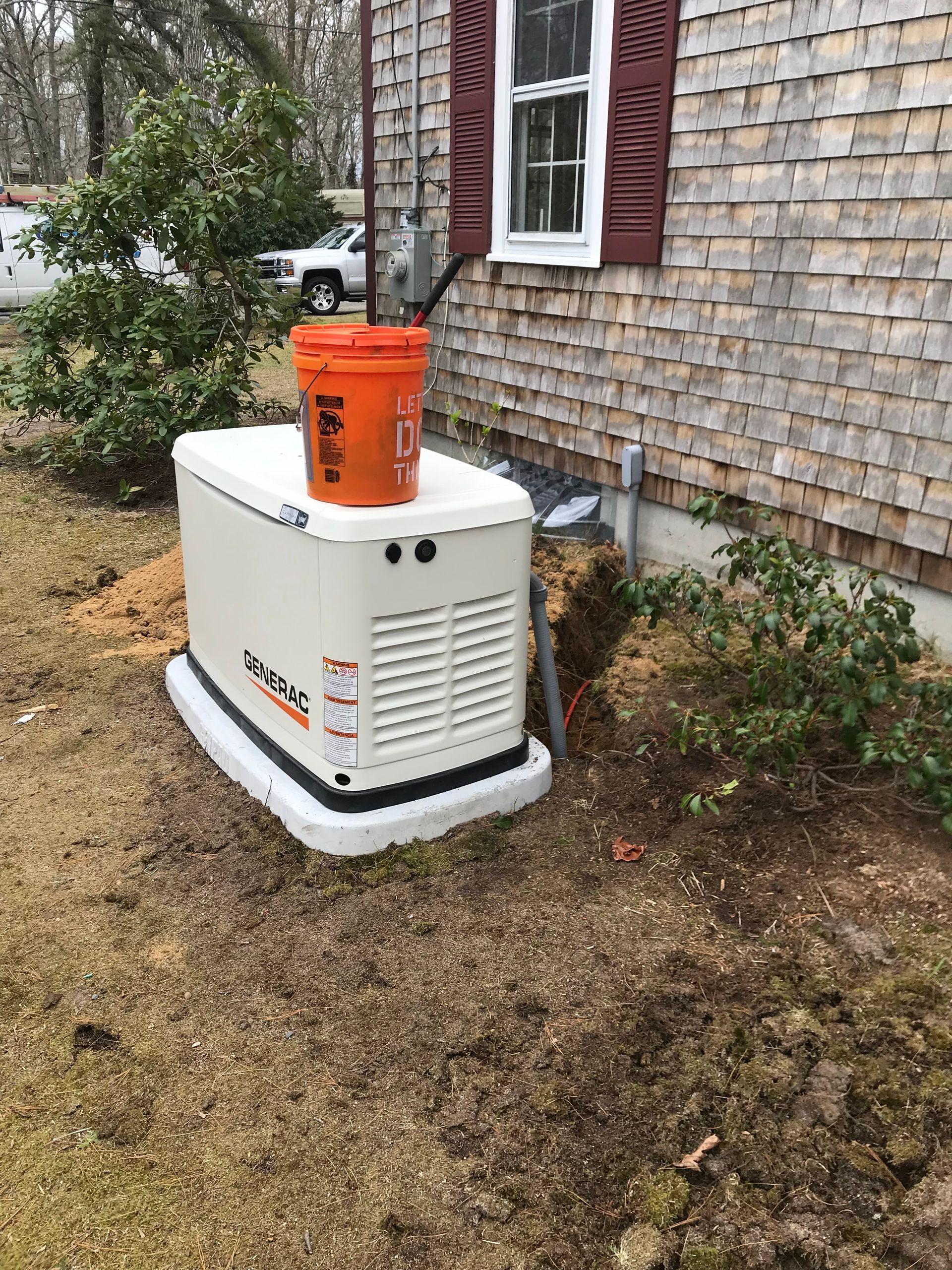 White generator outside a brick house with an orange bucket on top, beside shrubs and grass.