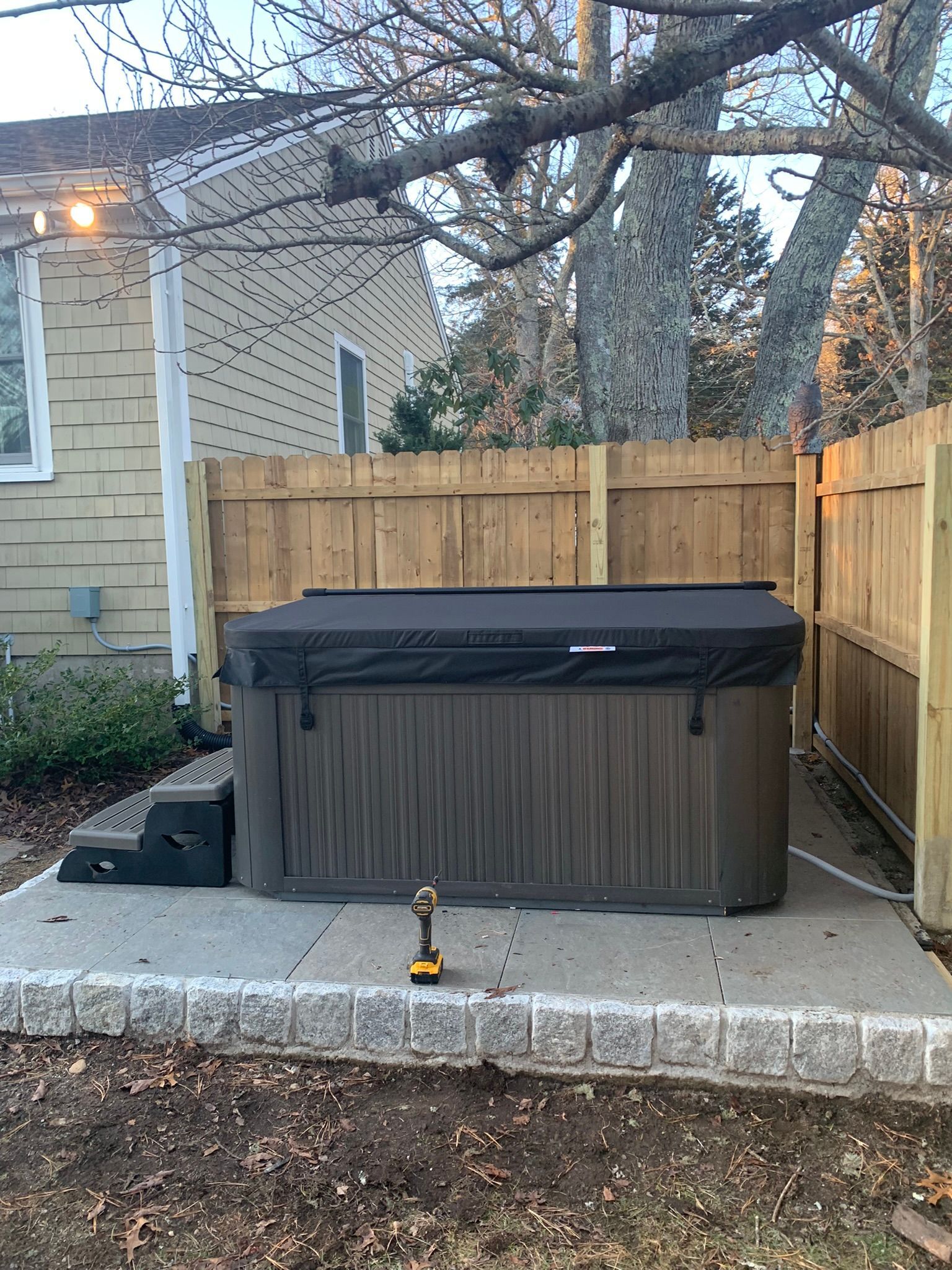 Covered hot tub in a fenced backyard beside a stone patio, with a small tool in front.