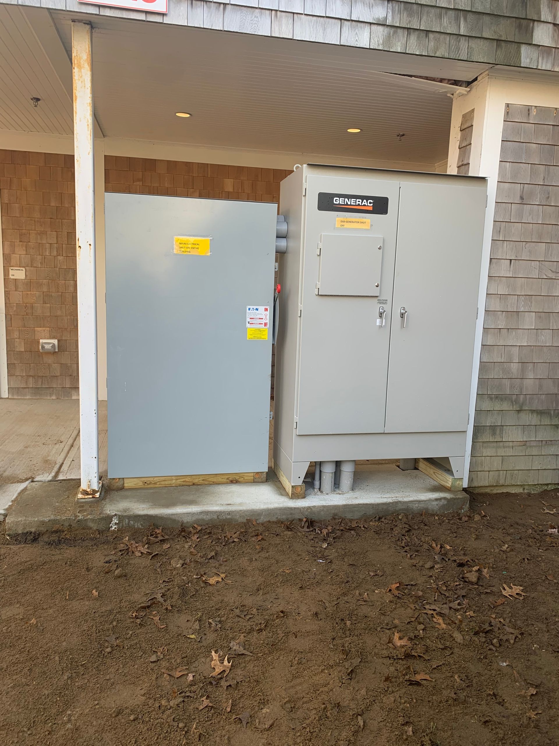Two gray electrical cabinets under a covered outdoor area, beside brick walls and a dirt ground.