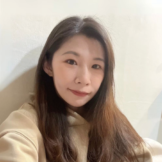 一位長著黑髮、穿著米色上衣的人,對著白牆露出溫柔的微笑。