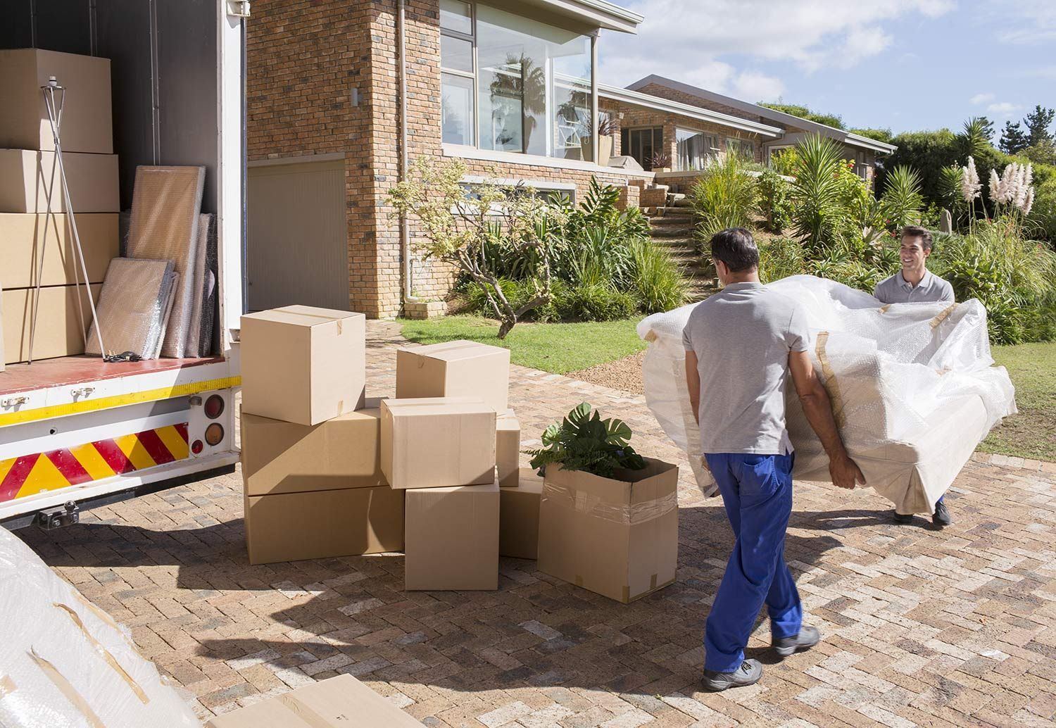'Two movers carrying wrapped furniture beside stacked boxes