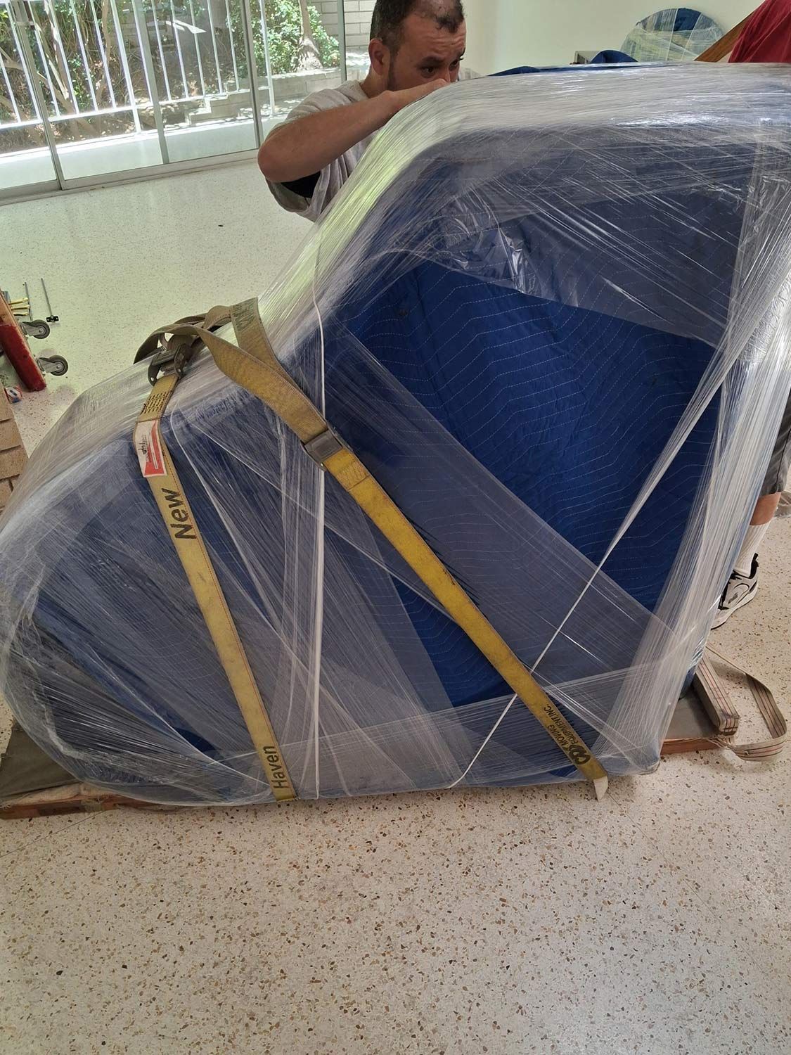 Blue seat wrapped in plastic and strapped with yellow tie-downs on a floor