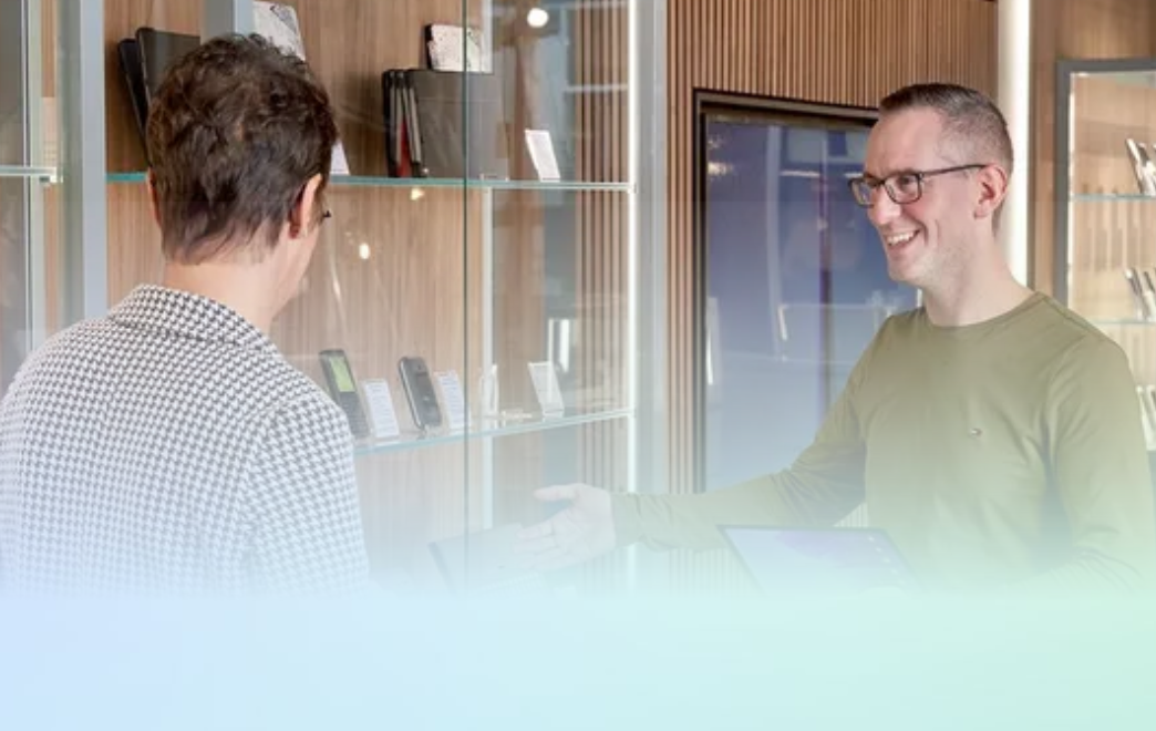 Twee mensen praten in een licht kantoor, een van hen glimlacht bij een glazen wand.