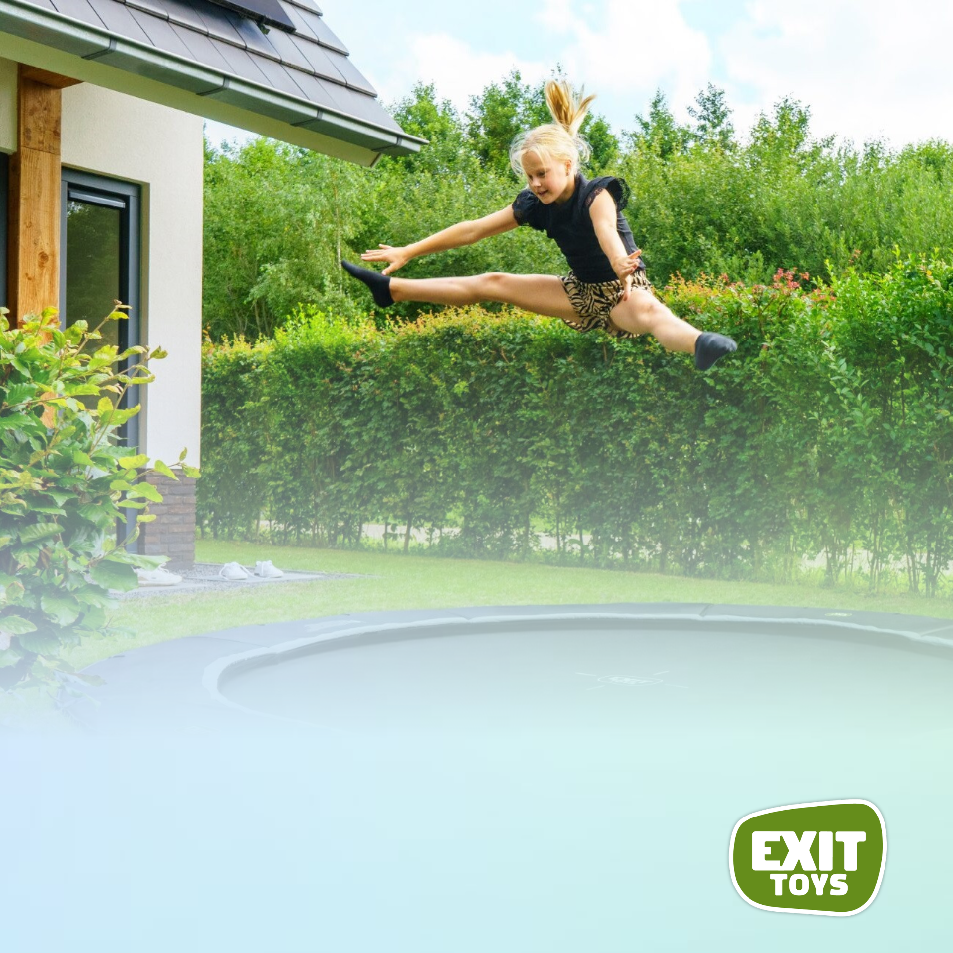 Een persoon maakt een spagaatsprong over een trampoline in de achtertuin, met een huis en een heg op de achtergrond.