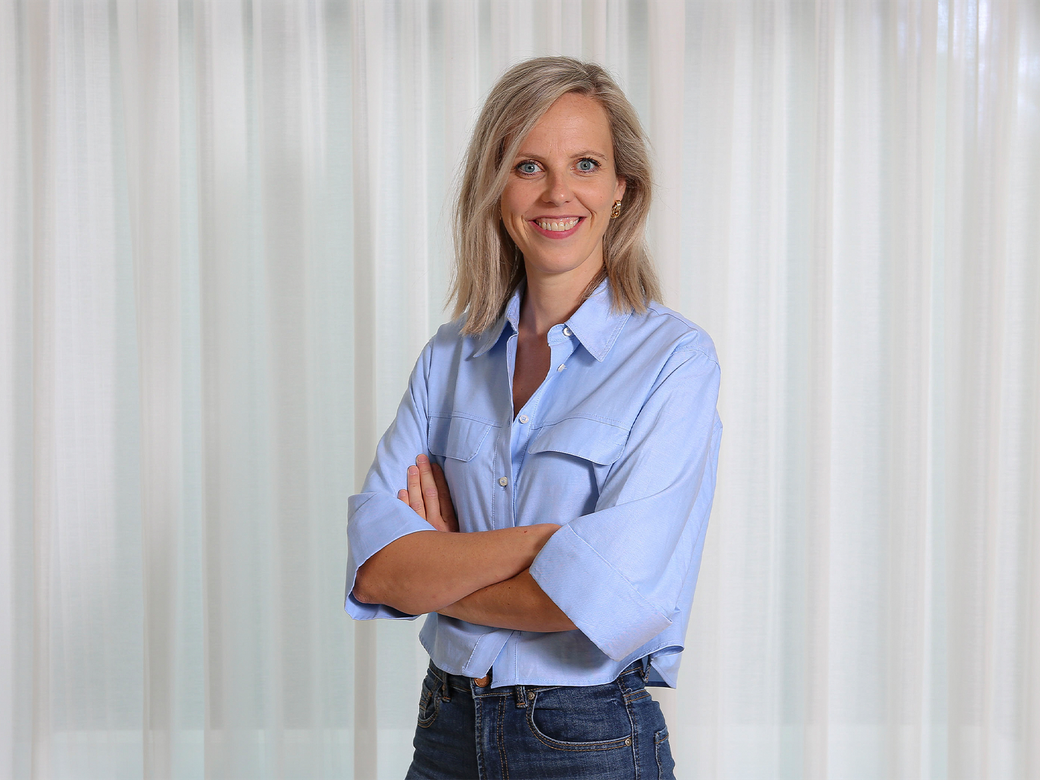Een lachende vrouw in een lichtblauwe blouse met de armen over elkaar, staand voor witte gordijnen.