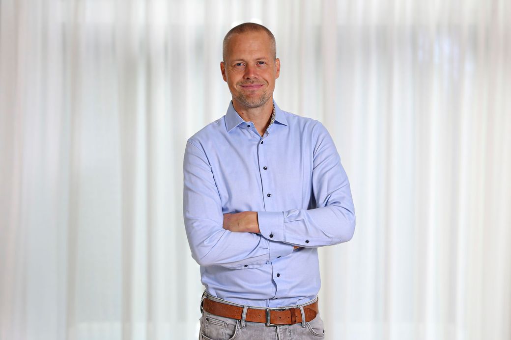 Een man staat met gekruiste armen in een lichtblauw shirt tegen een witte gordijnachtergrond.