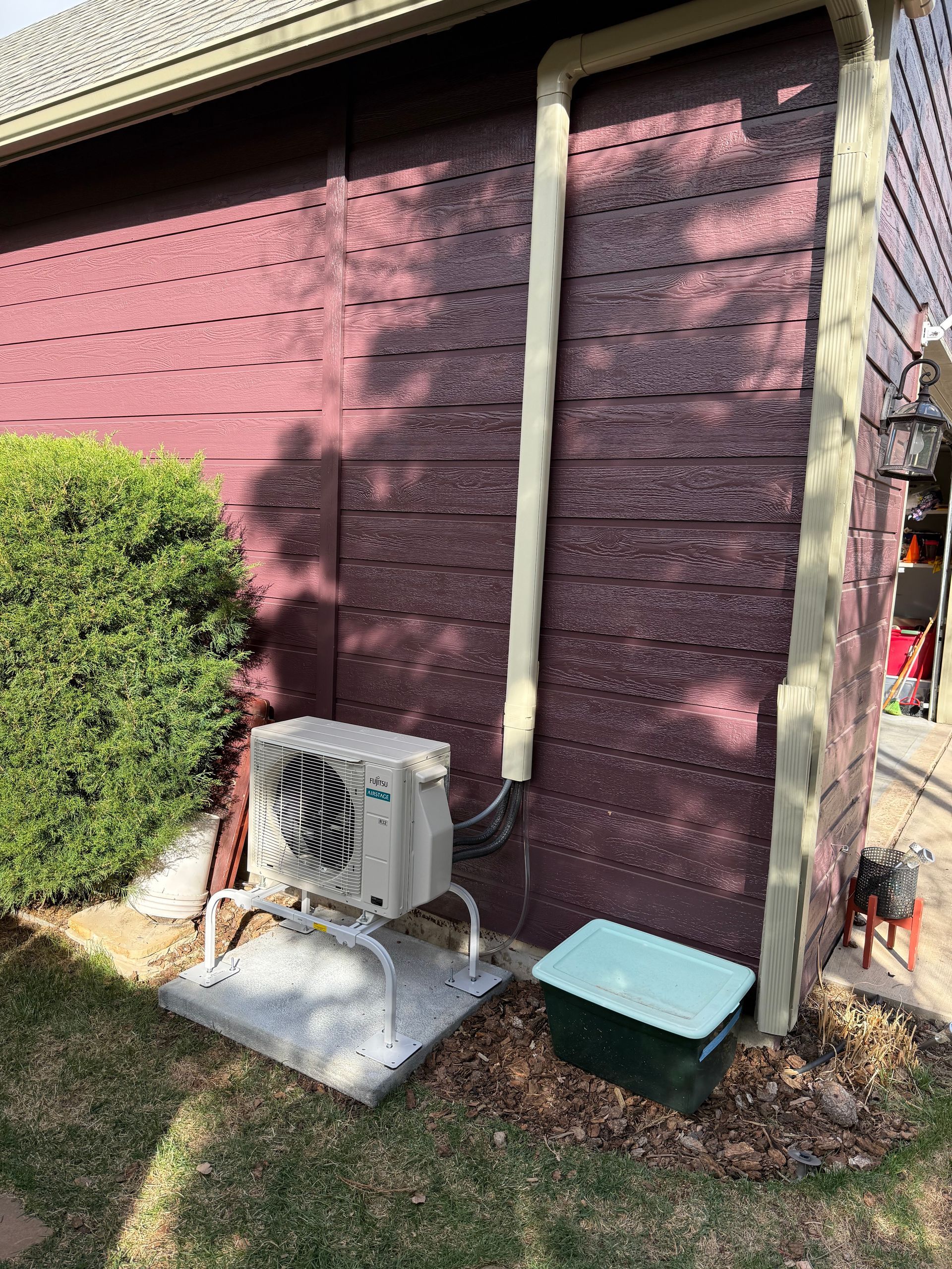 Exterior of a maroon house wall with an outdoor AC unit, small hedge, and green storage box on the lawn.