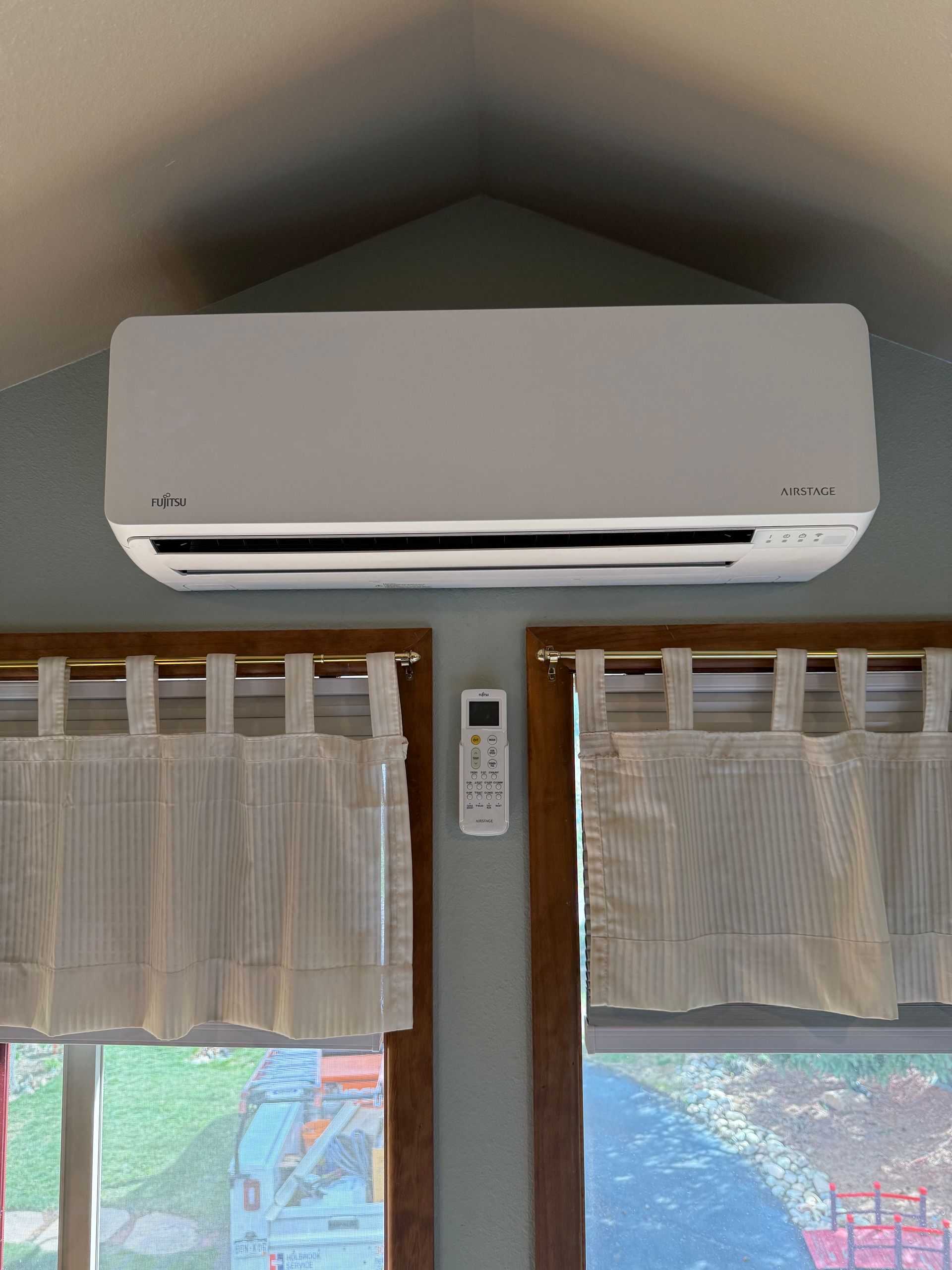 White wall-mounted air conditioner above windows with curtains and a remote on the wall
