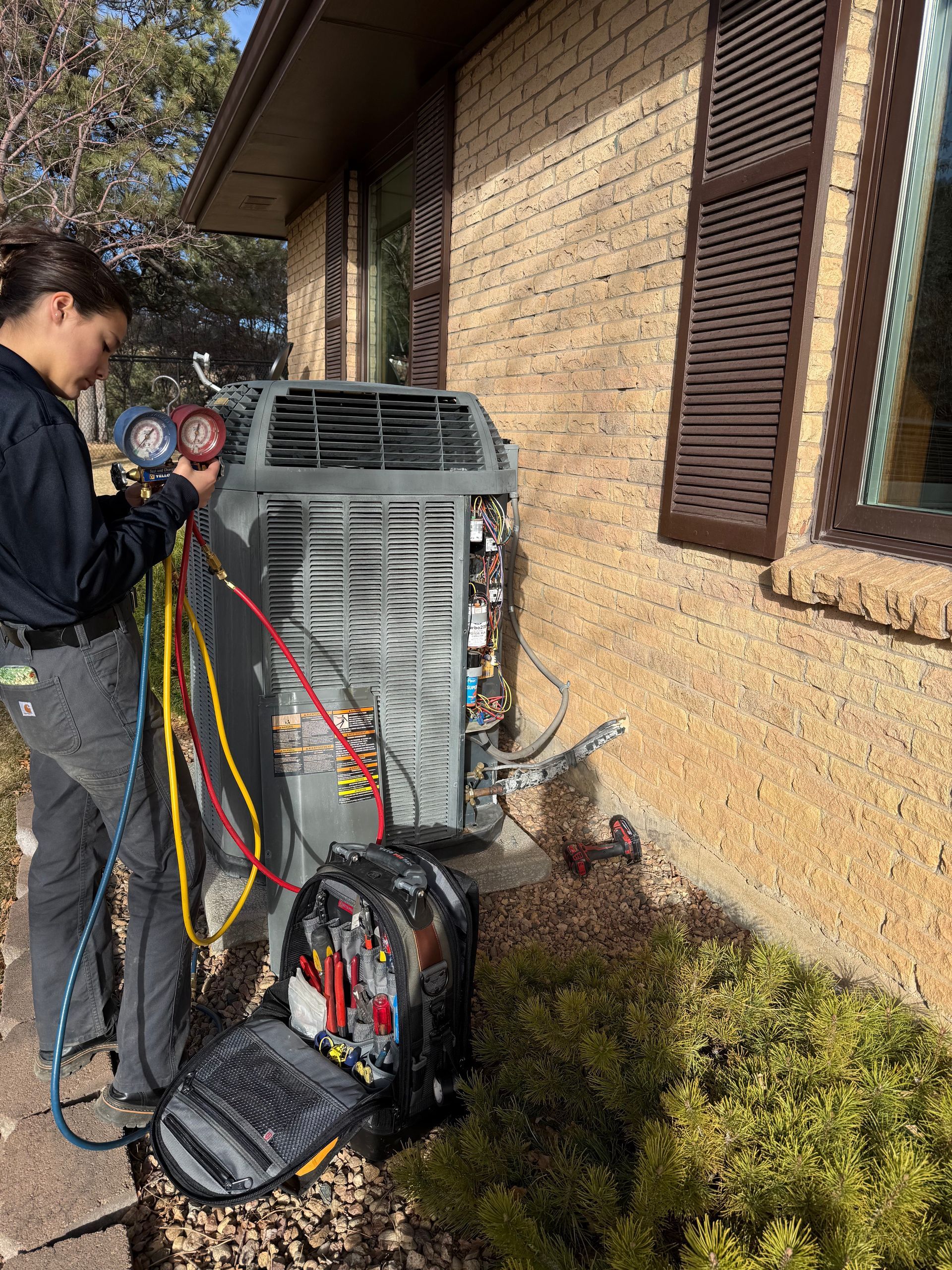 Technician servicing outdoor HVAC unit beside a brick house, with gauges and tools attached