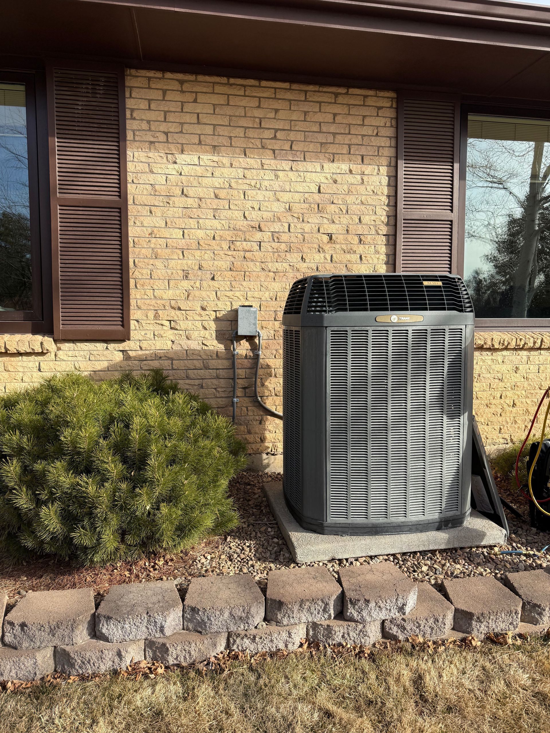 Outdoor HVAC unit beside a brick house, with shrubs and stone edging in front of shuttered windows