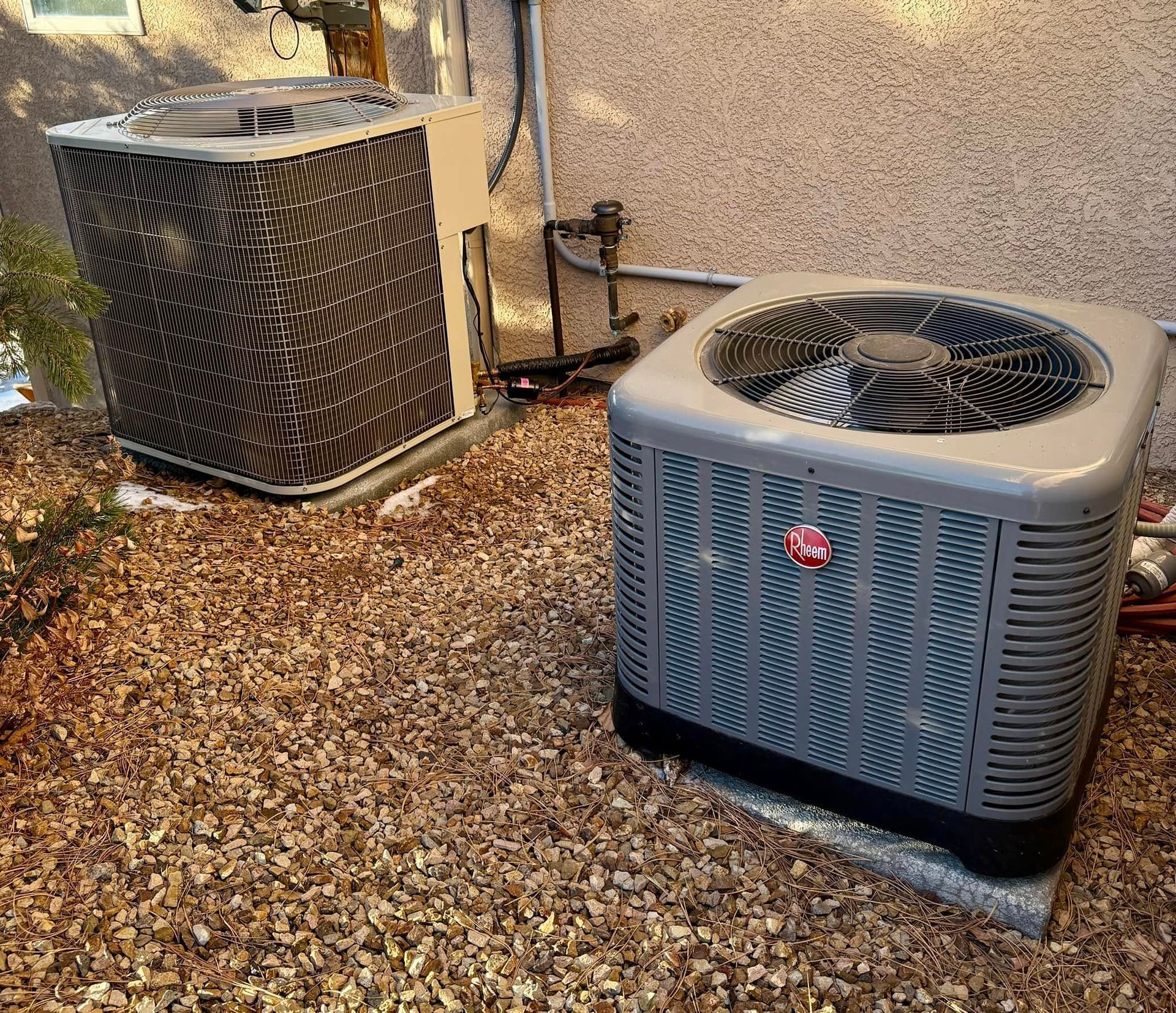 Two outdoor air conditioning units beside a stucco wall on gravel in a yard