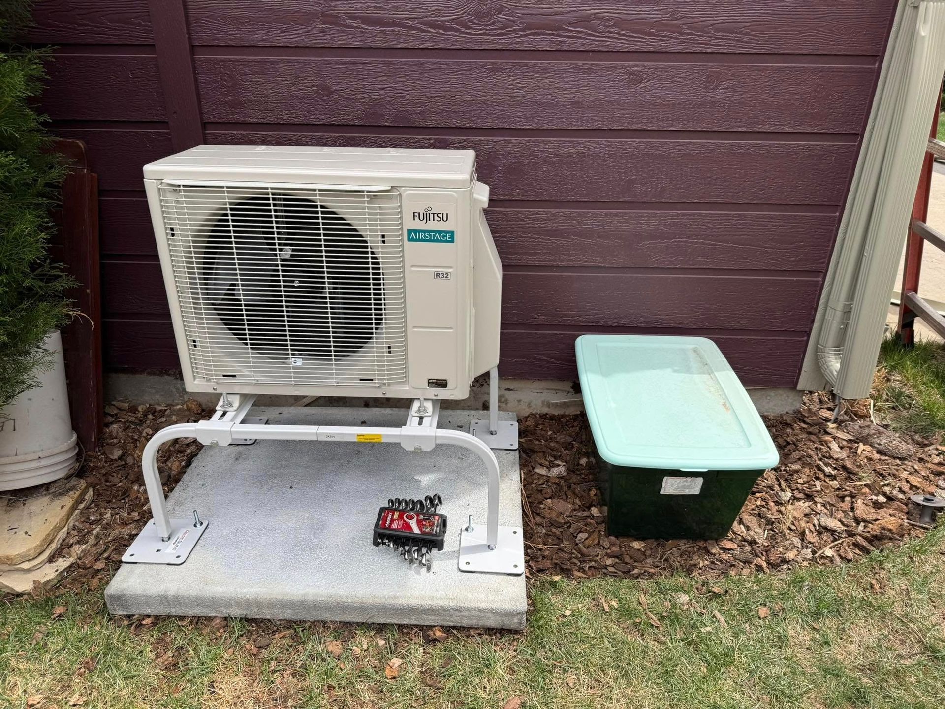 Outdoor AC unit on a concrete pad beside a brown house wall, with a green storage bin nearby.