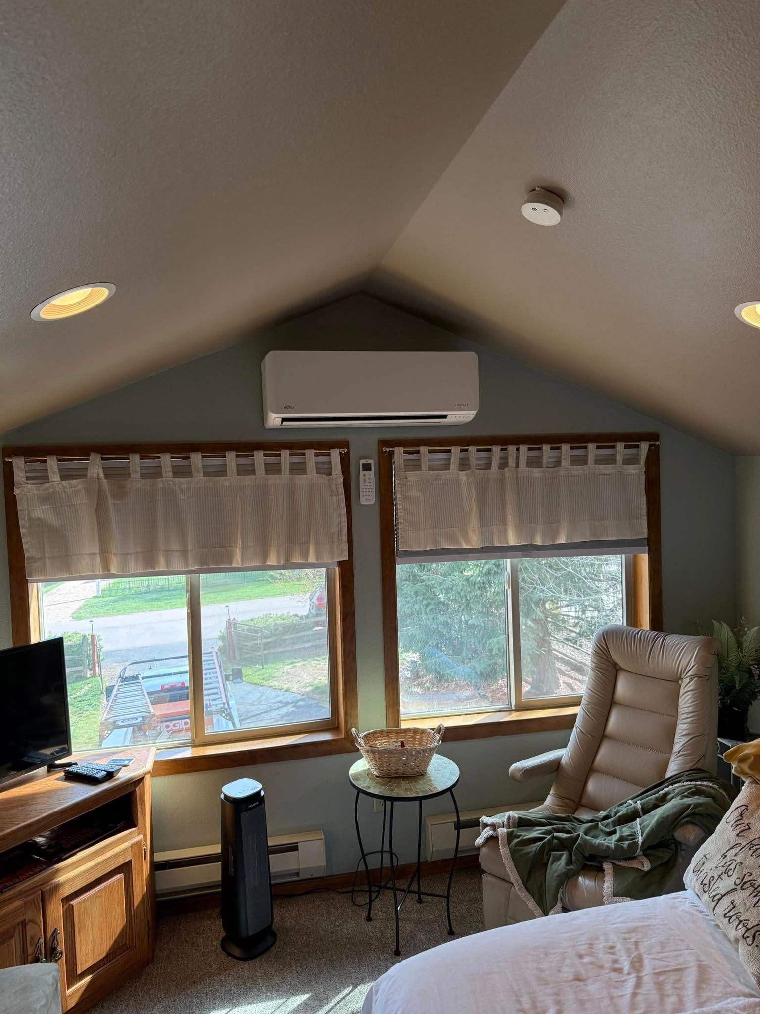 Cozy living room with large windows, wood trim, armchair, TV, and an air conditioner on a sloped ceiling.