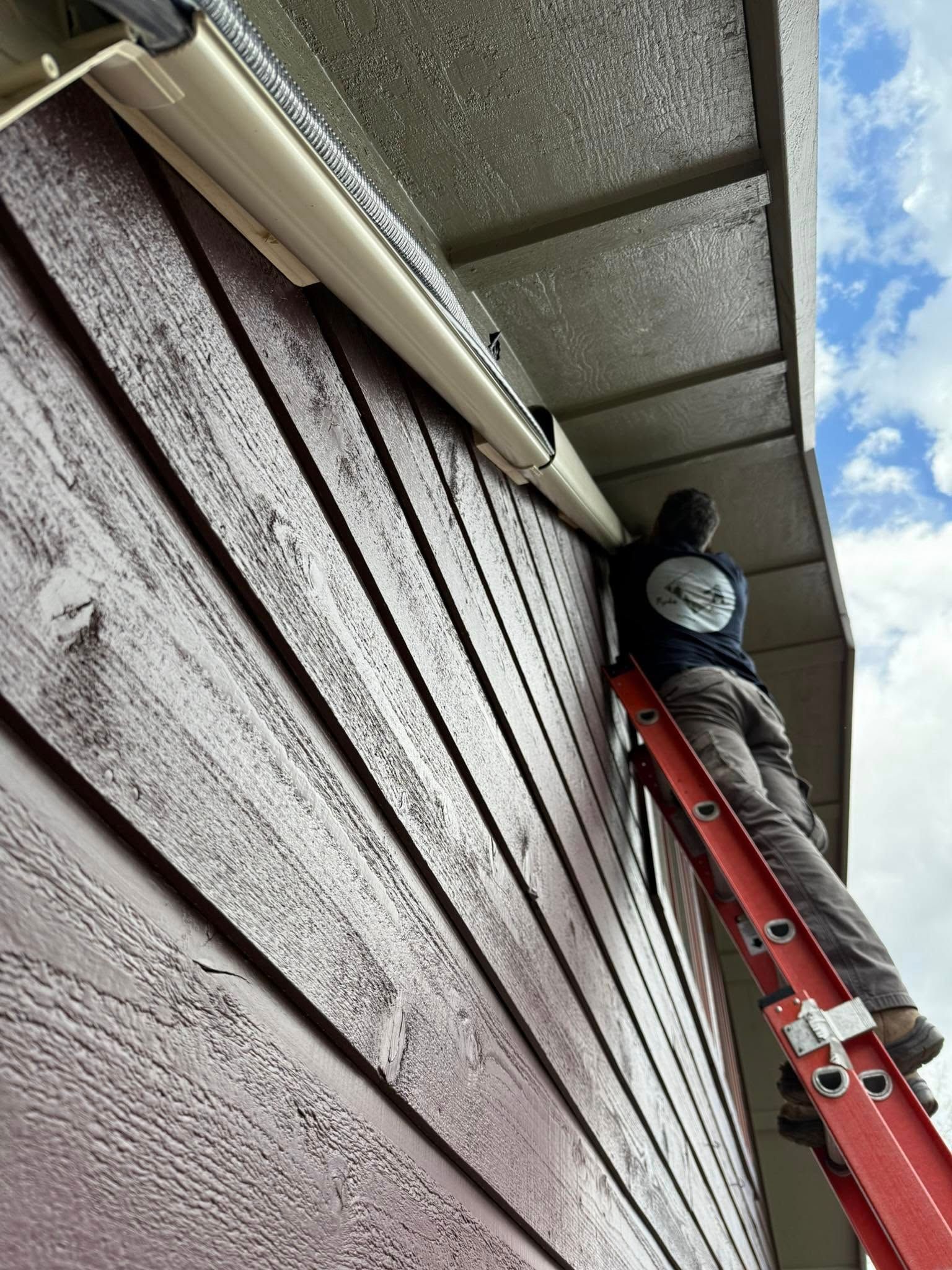 Worker on a red ladder cleaning a house exterior under the roof eaves
